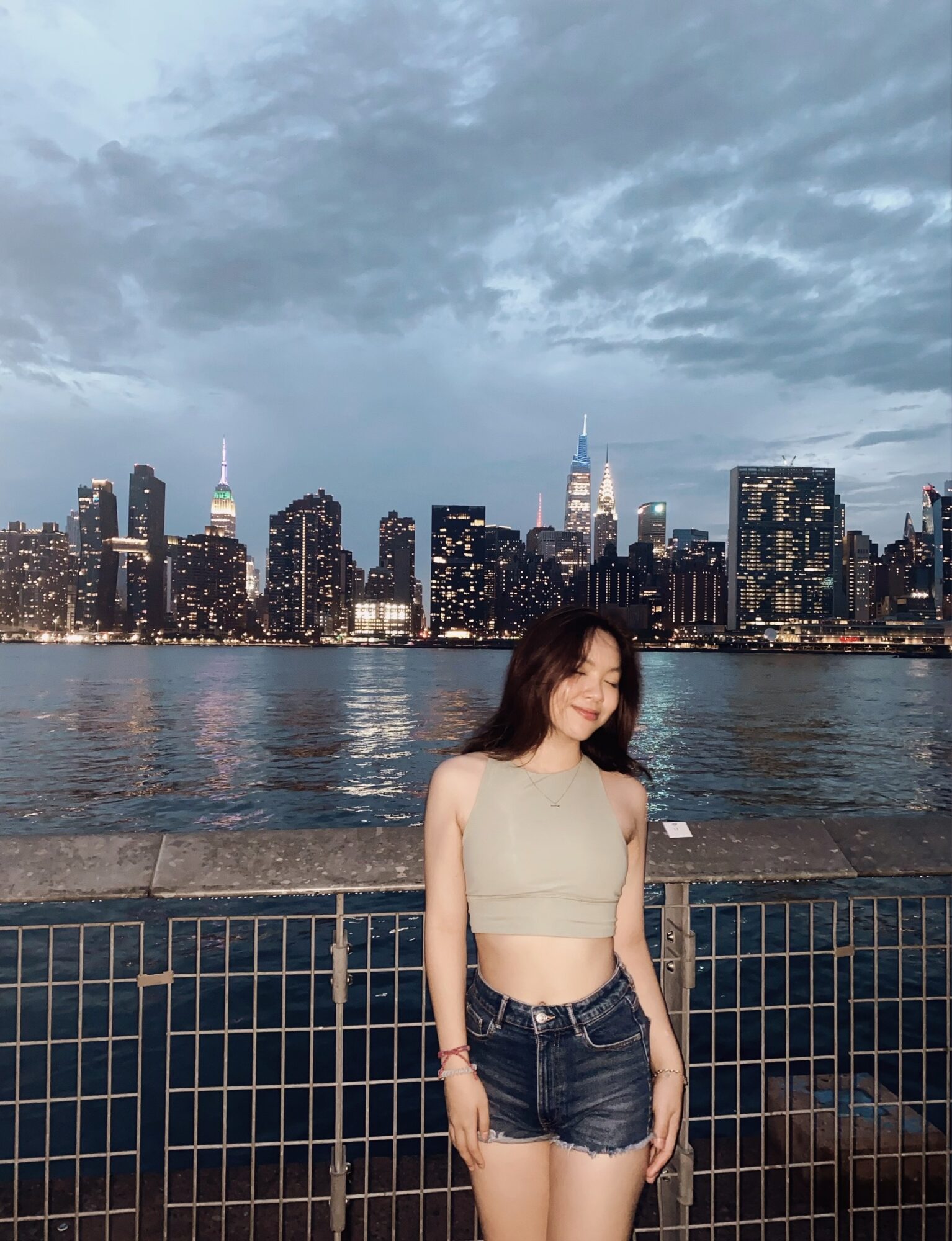 Young woman with dark hair in a sleeveless crop top and denim shorts standing by a railing with city skyline and water behind her