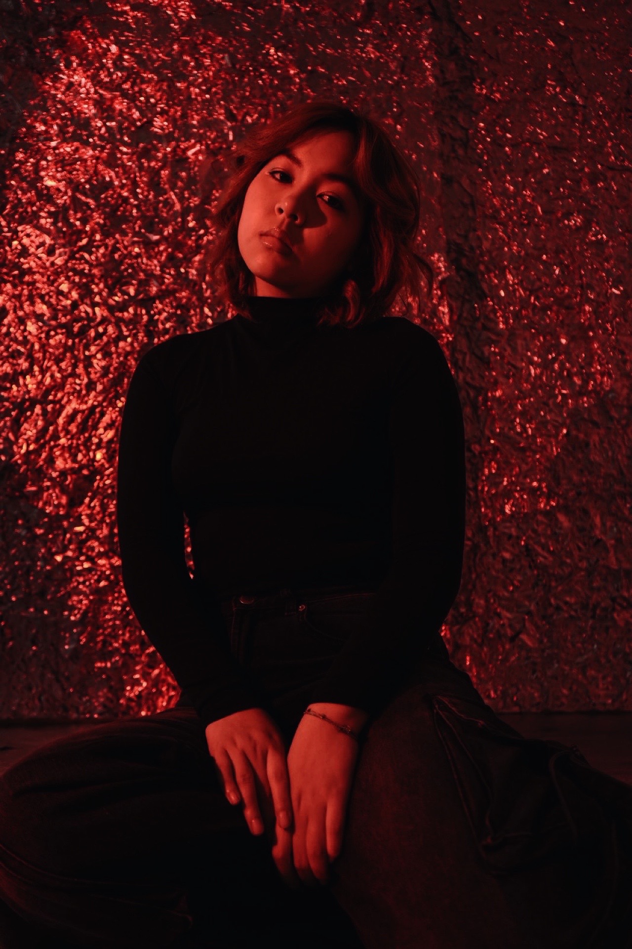 Person with shoulder-length hair sitting against a textured red background, wearing a black top, looking at the camera.