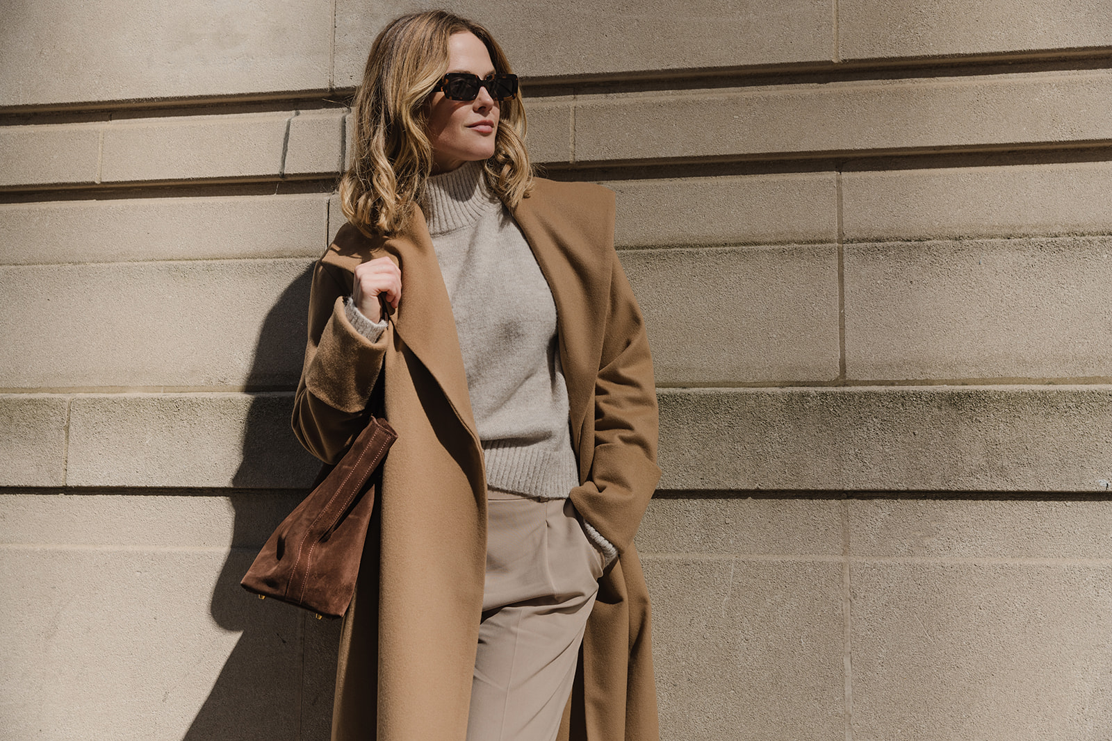 Woman with sunglasses in a tan coat and gray sweater standing against a wall, holding a bag.