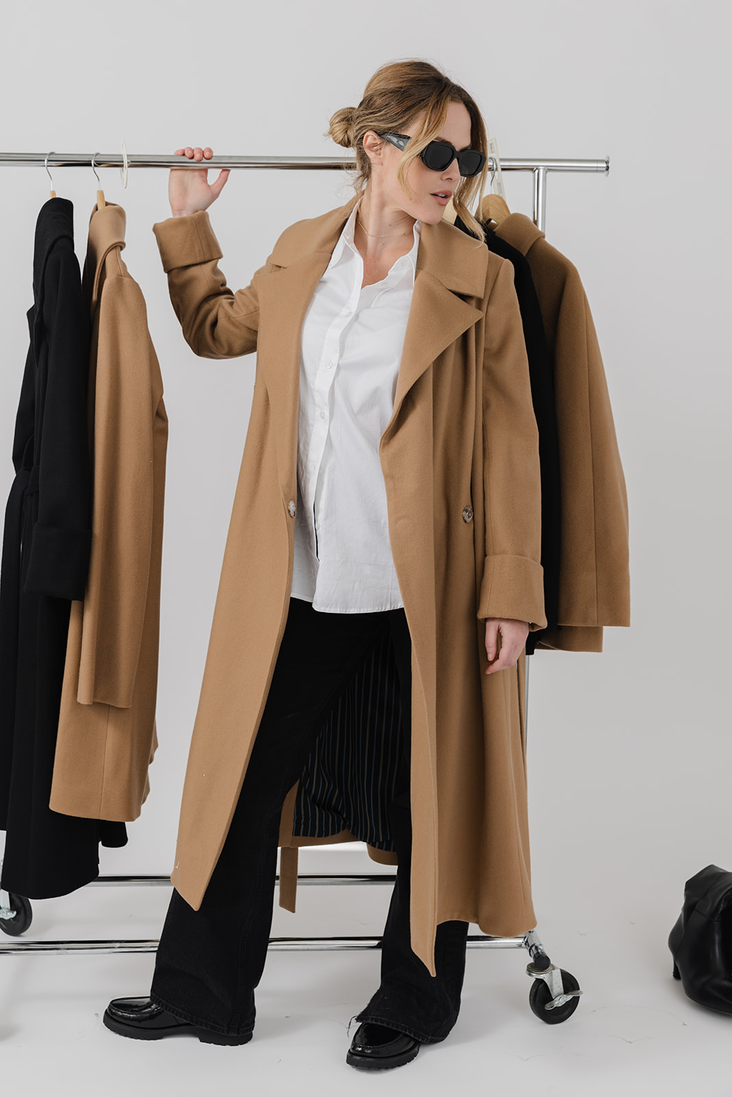 Woman with sunglasses wearing a long tan coat, white shirt, and black pants, standing next to a clothing rack with coats.