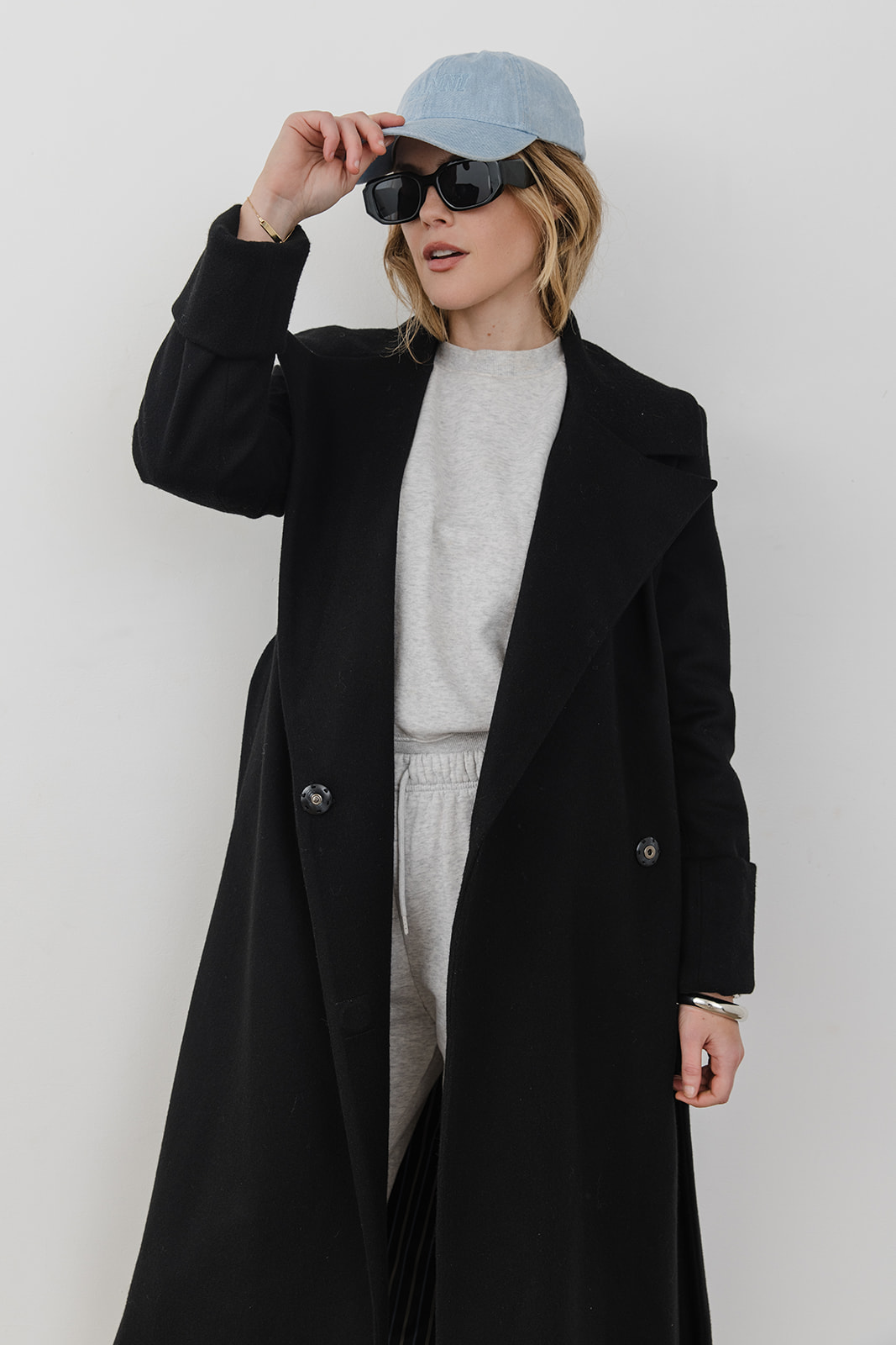 Woman wearing sunglasses, a light blue cap, black coat, and gray outfit, adjusting her cap against a plain white background.