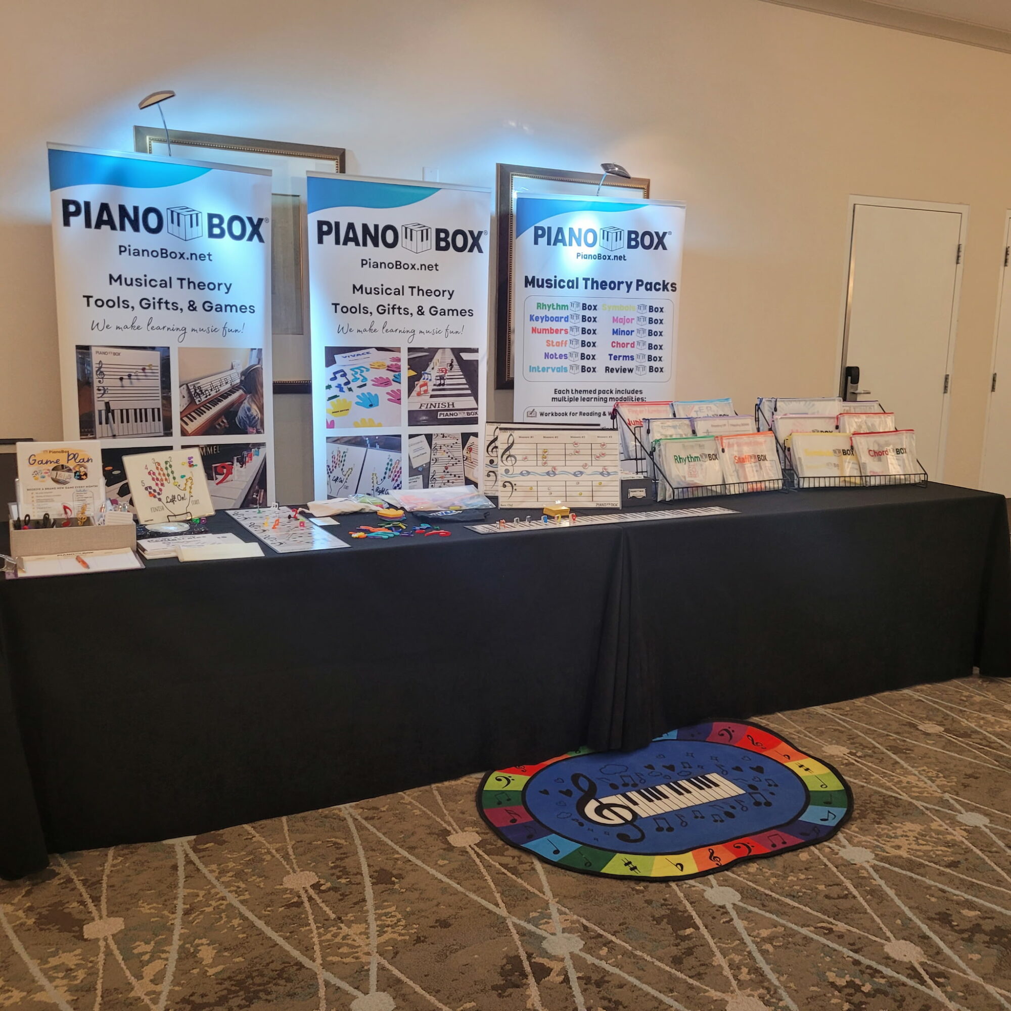 Table with music books and pamphlets, two display boards, colorful circular rug, and a beige wall with a door in the background.