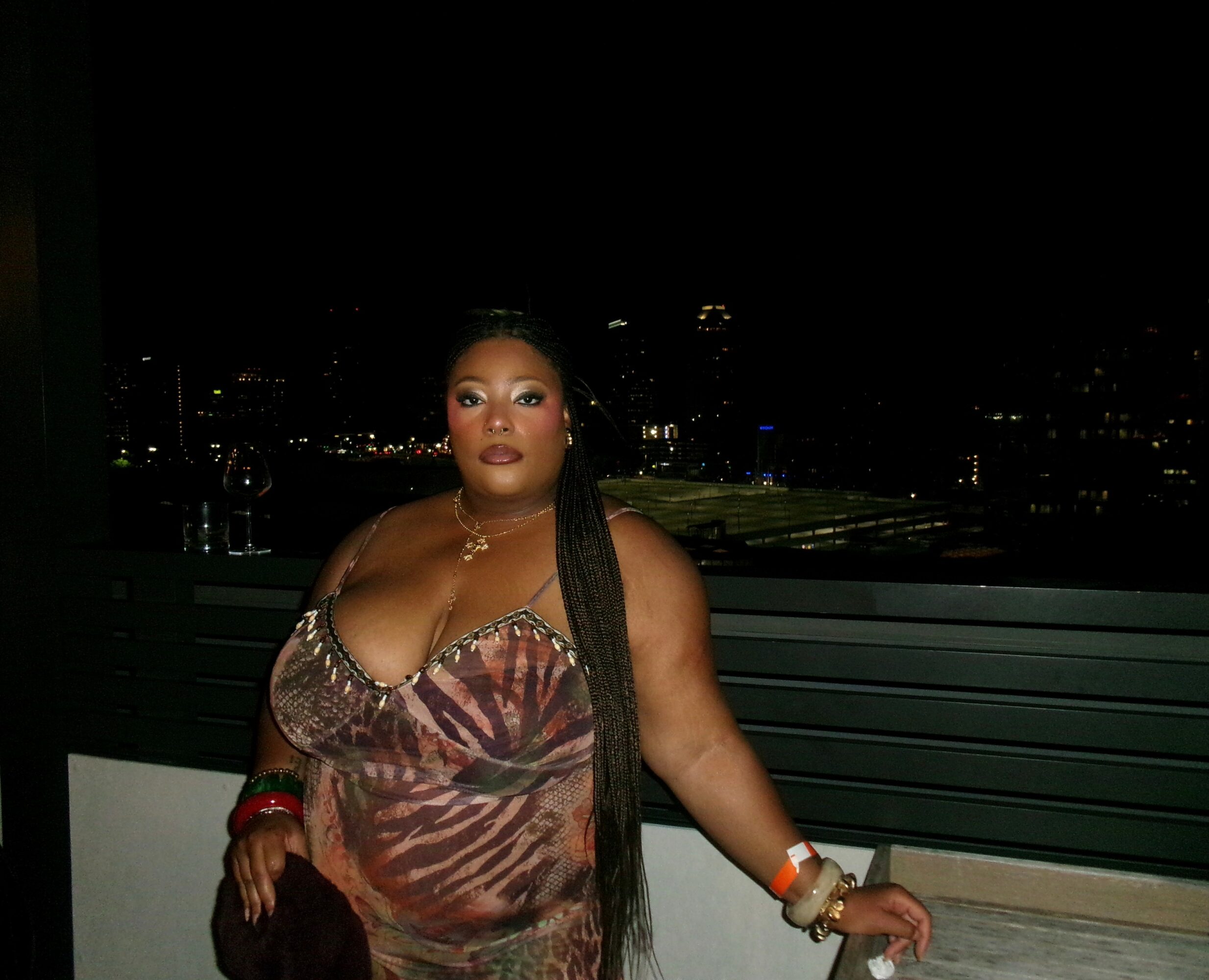 Woman with long braided hair in a patterned dress standing outdoors at night with city lights in background.