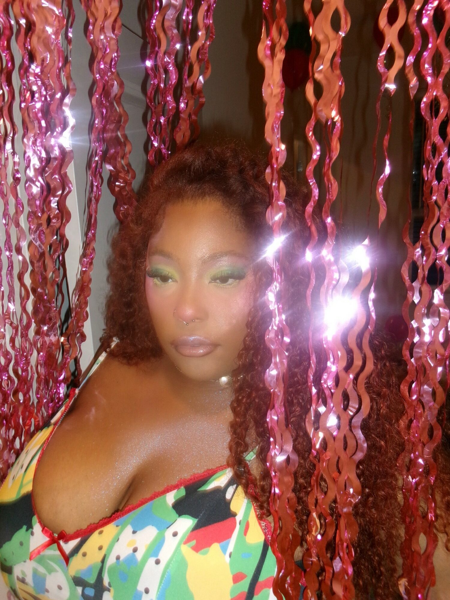 Woman with curly hair and colorful makeup, surrounded by pink and red hanging decorations, looking at camera.