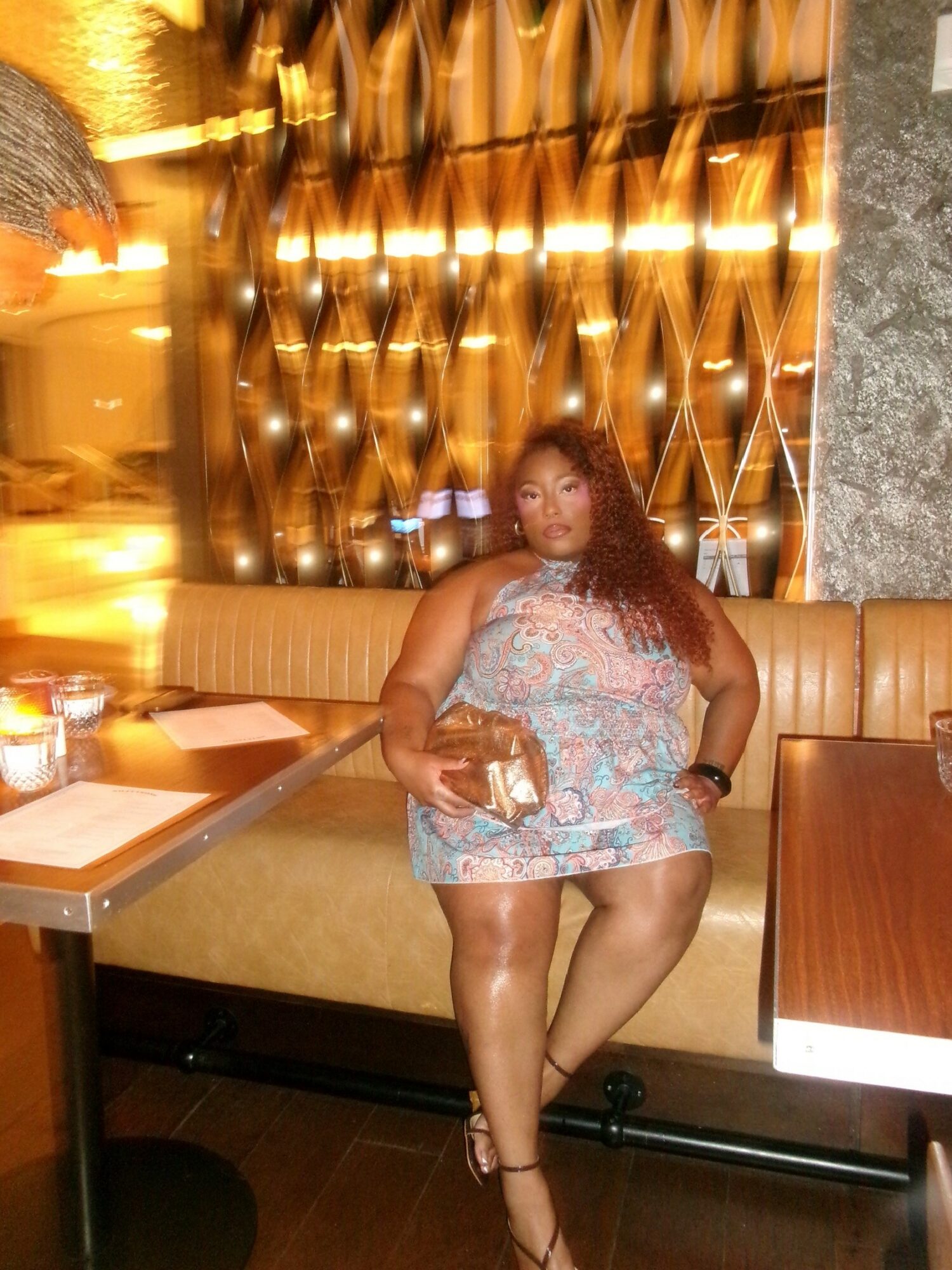 Woman in a patterned dress sitting on a booth in a restaurant, with a decorative wall behind her.
