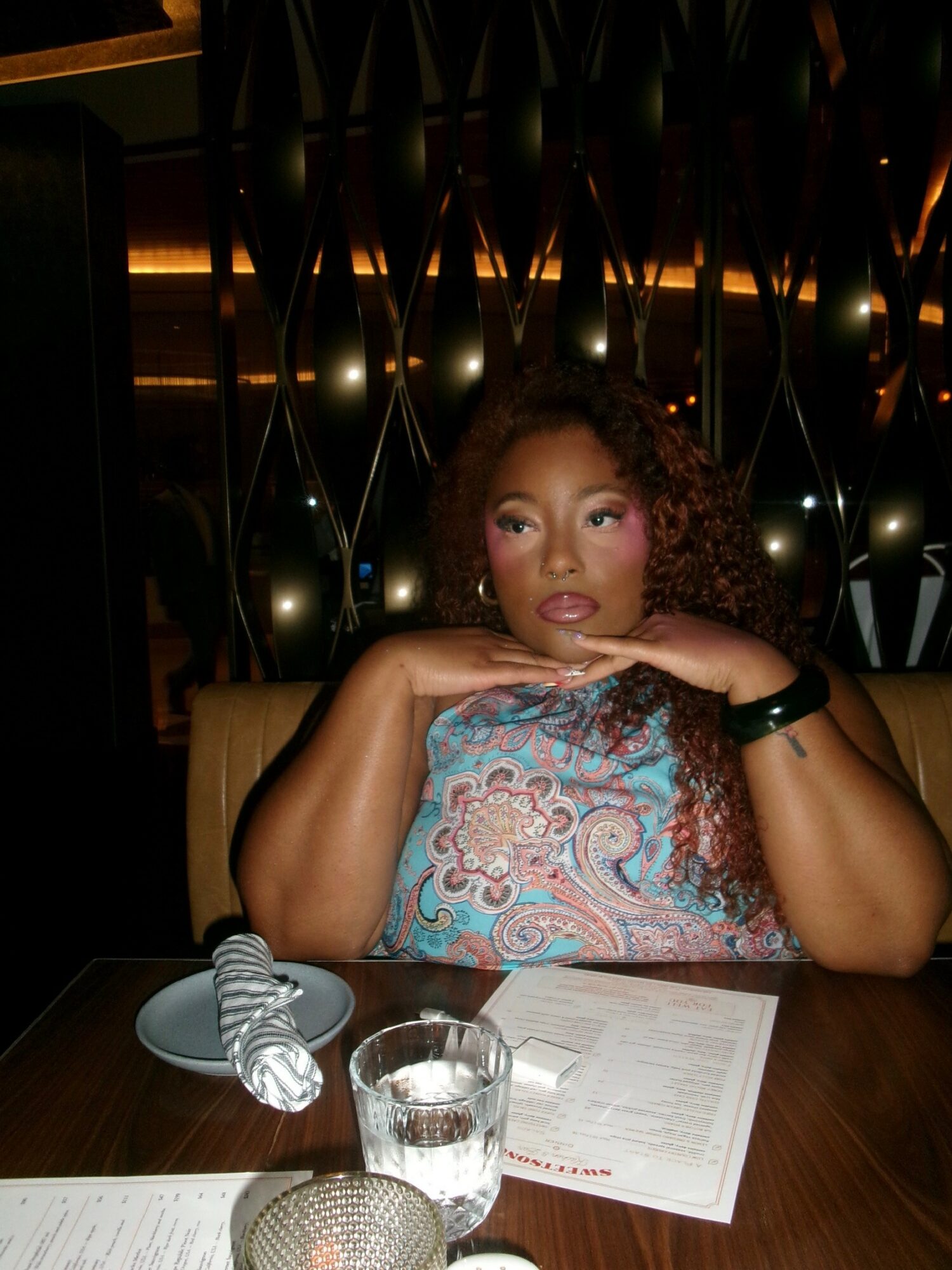 Woman with curly hair resting her chin on her hands at a table in a dimly lit setting.