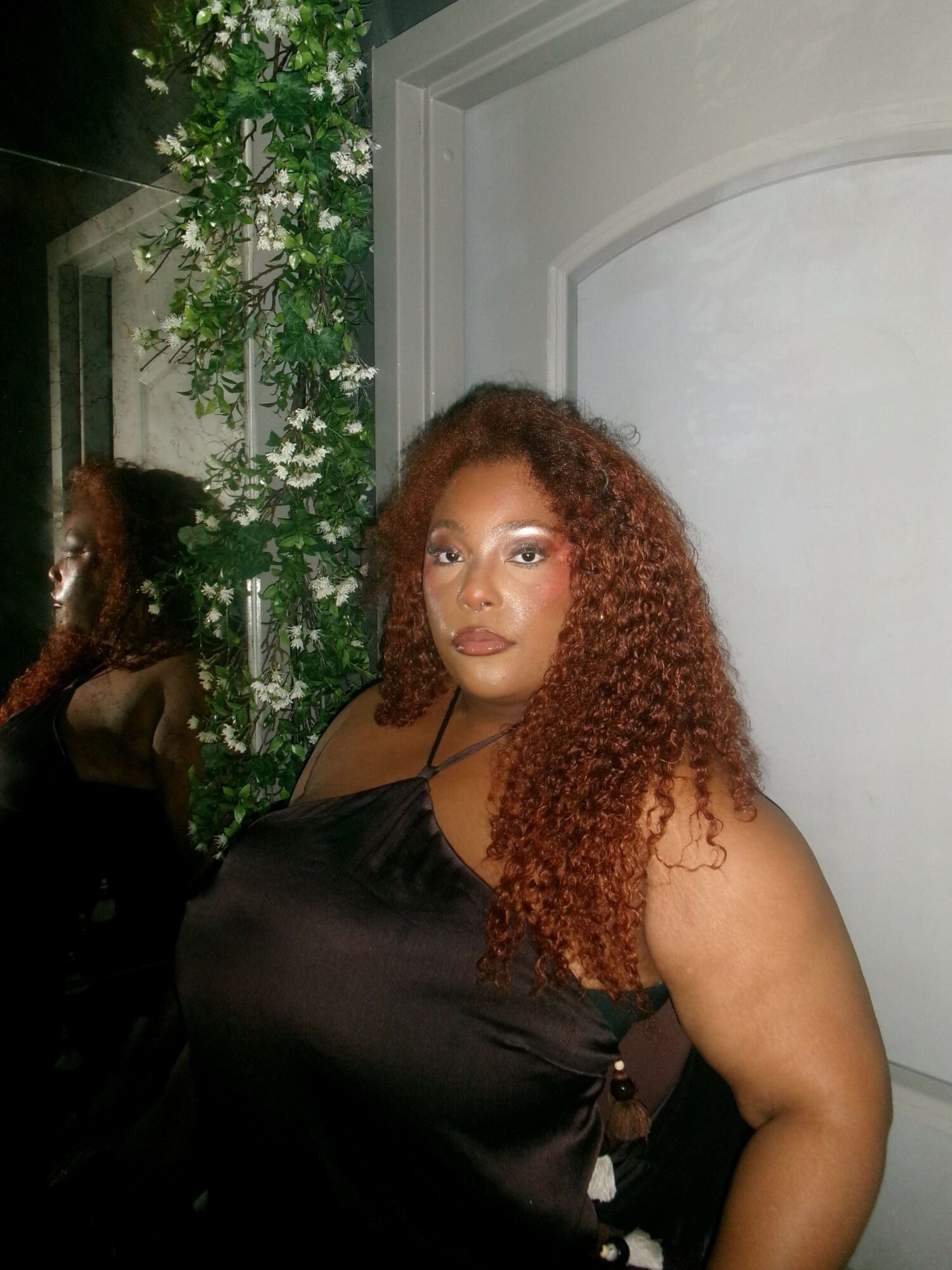 Woman with curly hair in a black top standing near a window with green plants, looking at the camera.