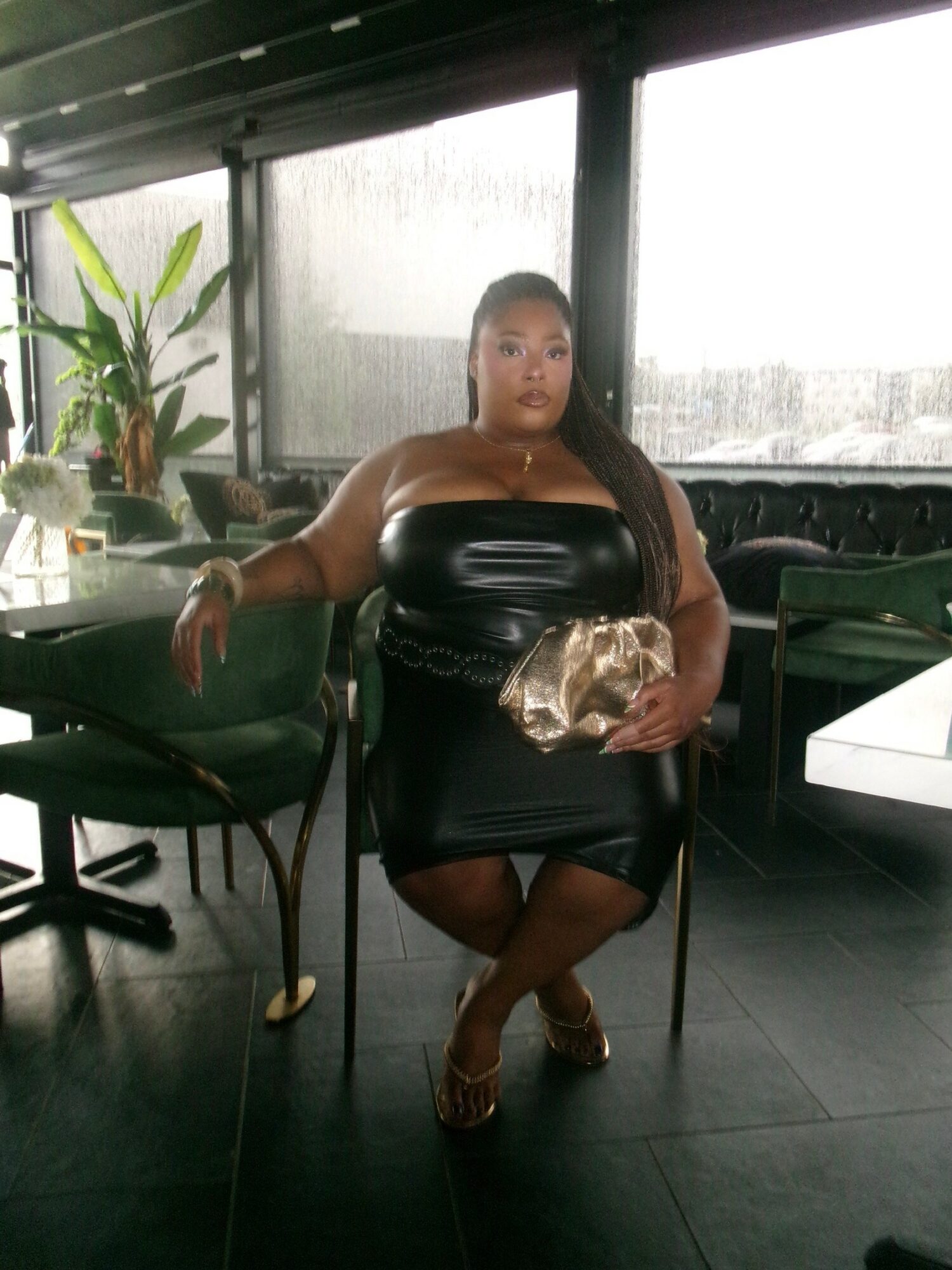 Woman in a black strapless dress sitting at a table holding a bag, with large windows and plants in the background.
