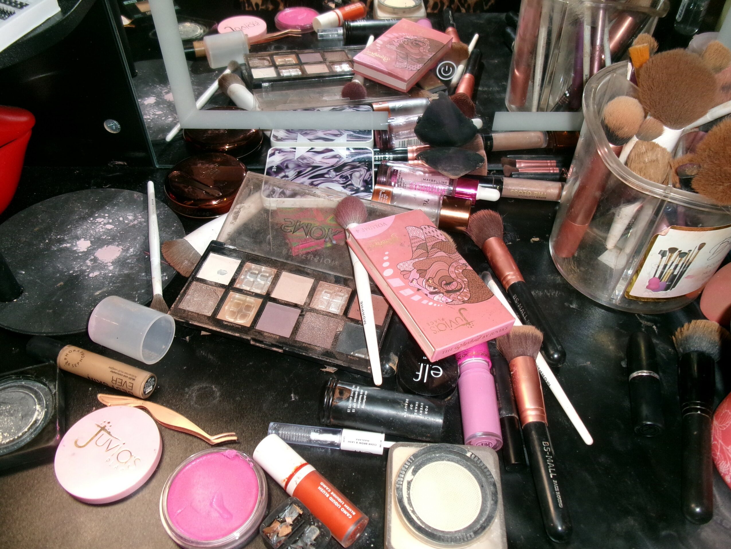 Makeup and cosmetic products scattered on a black surface, including eyeshadow palettes, lipsticks, blush, and brushes.