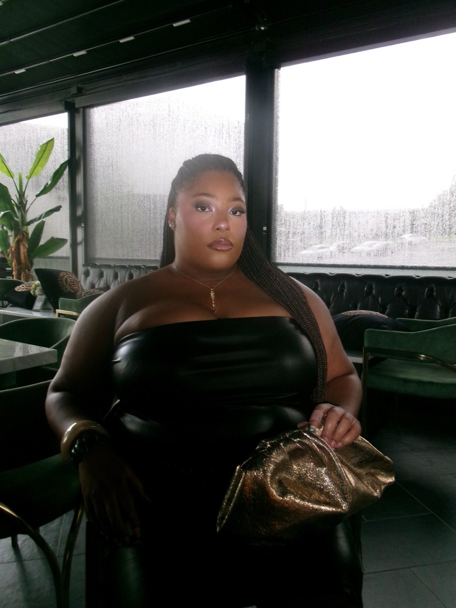 Woman sitting indoors near window with rain outside, holding a gold purse, wearing a strapless black top, looking at camera.