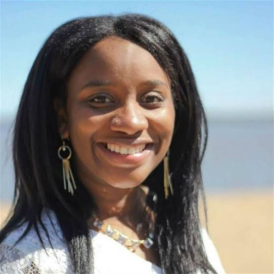 Woman smiling outdoors with beach and sky in background, wearing earrings and a necklace.