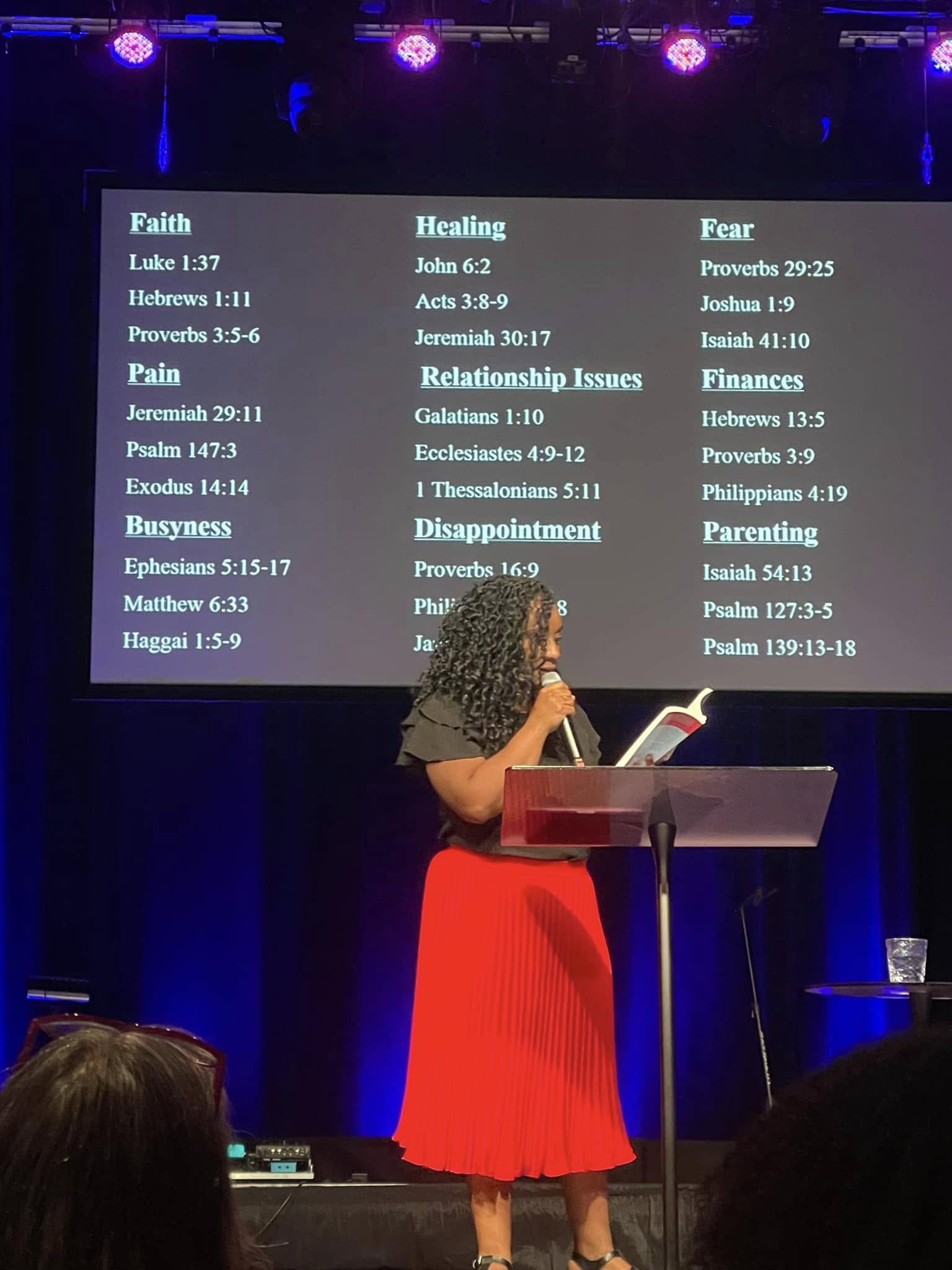 Woman in red dress speaking at a podium on stage with a large screen displaying words related to faith, healing, fear, and disappointment.