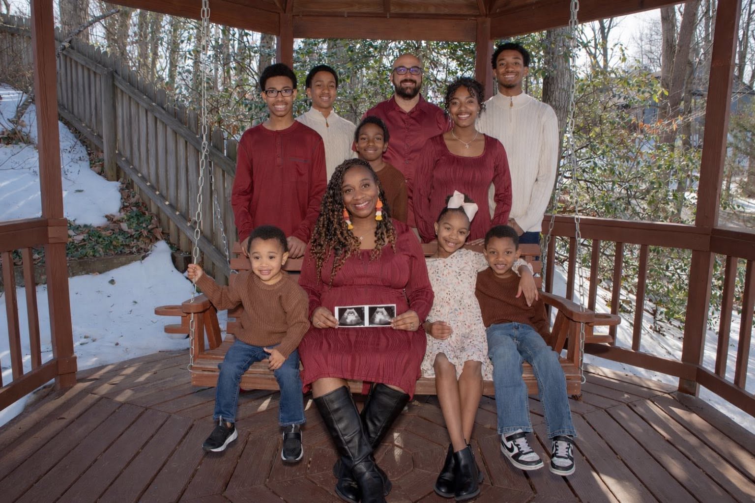 Group of nine people, including children and adults, sitting and standing on a wooden porch with trees and snow in background.