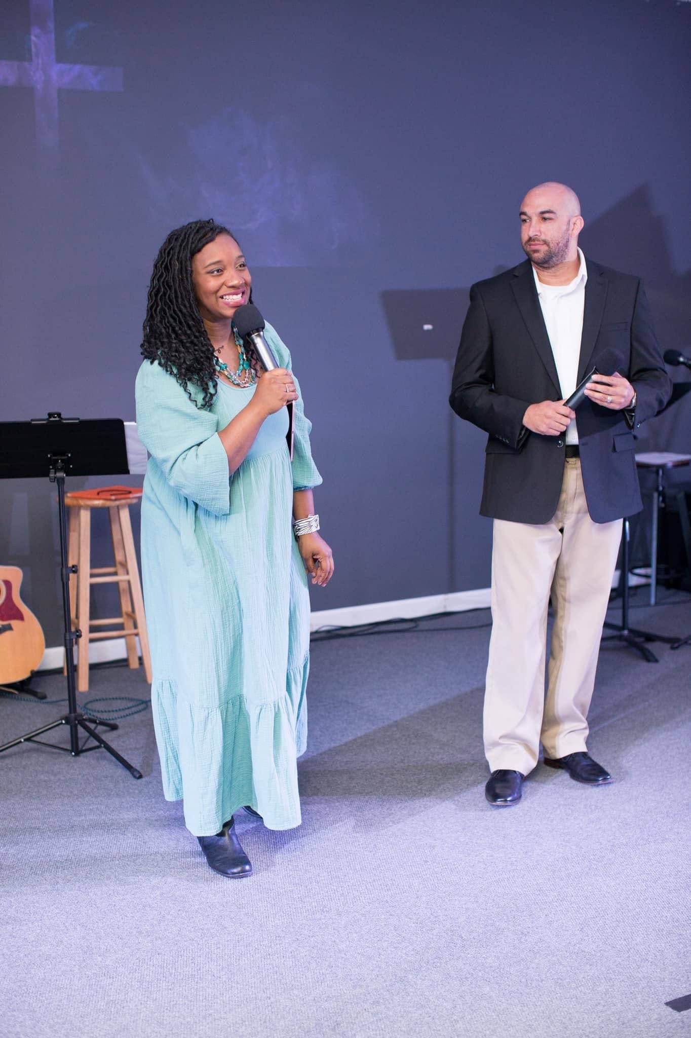 A woman and a man stand on stage, with musical instruments in the background, speaking to an audience.