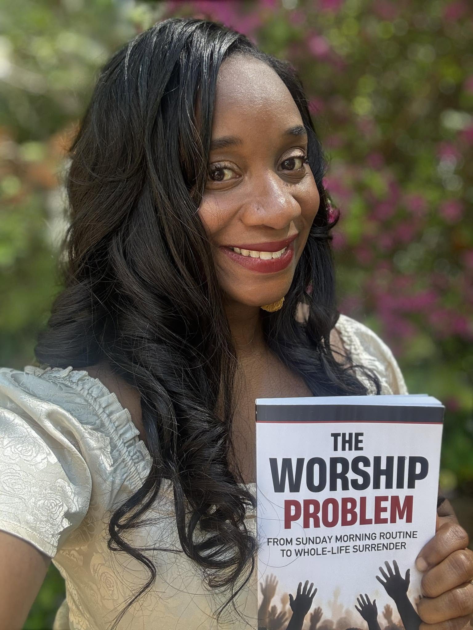 Smiling woman with long dark hair holding a book titled 'The Worship Problem' outdoors with pink flowers in background.