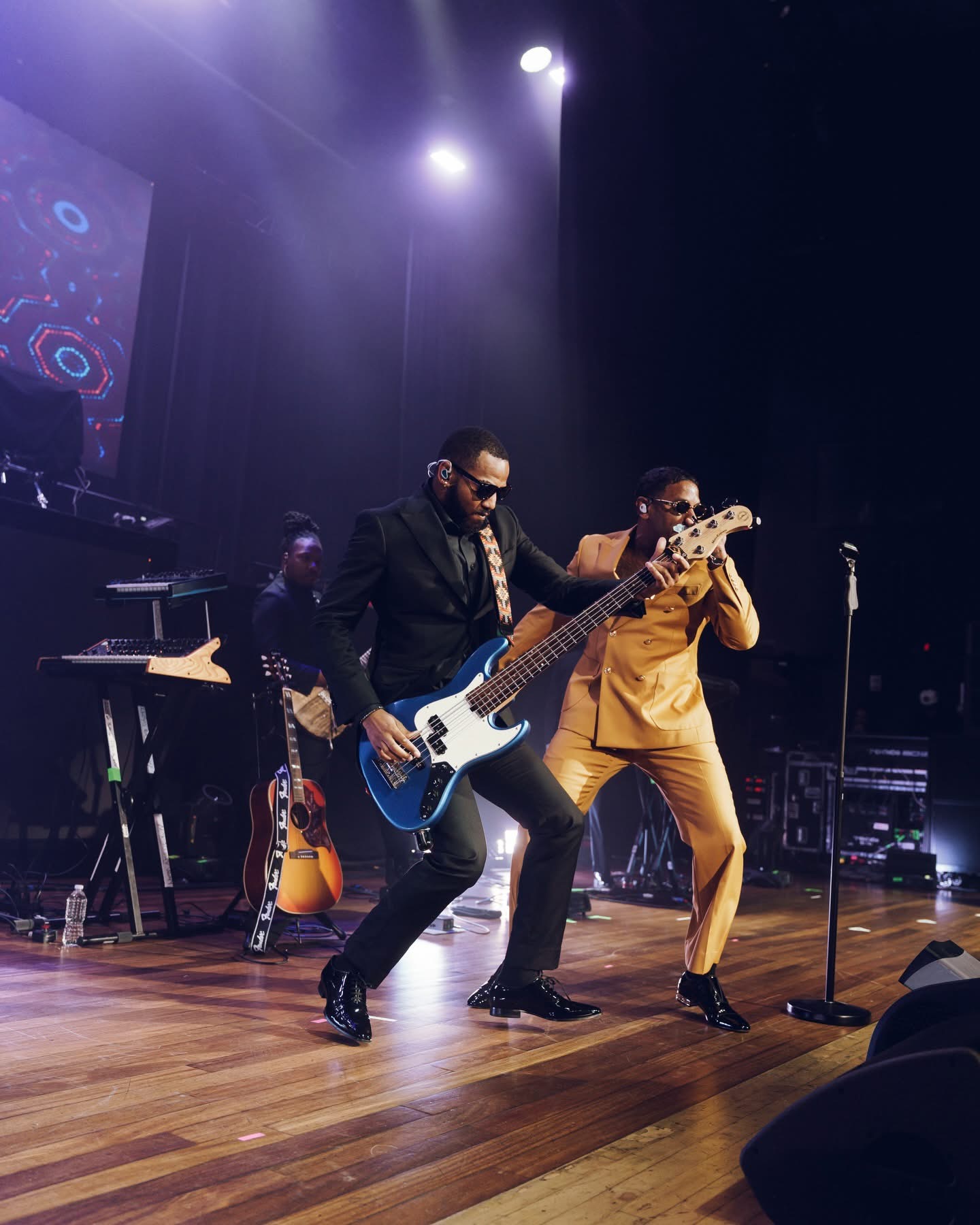 Two musicians perform on stage, one playing bass guitar and the other singing into a microphone, with stage lights overhead.
