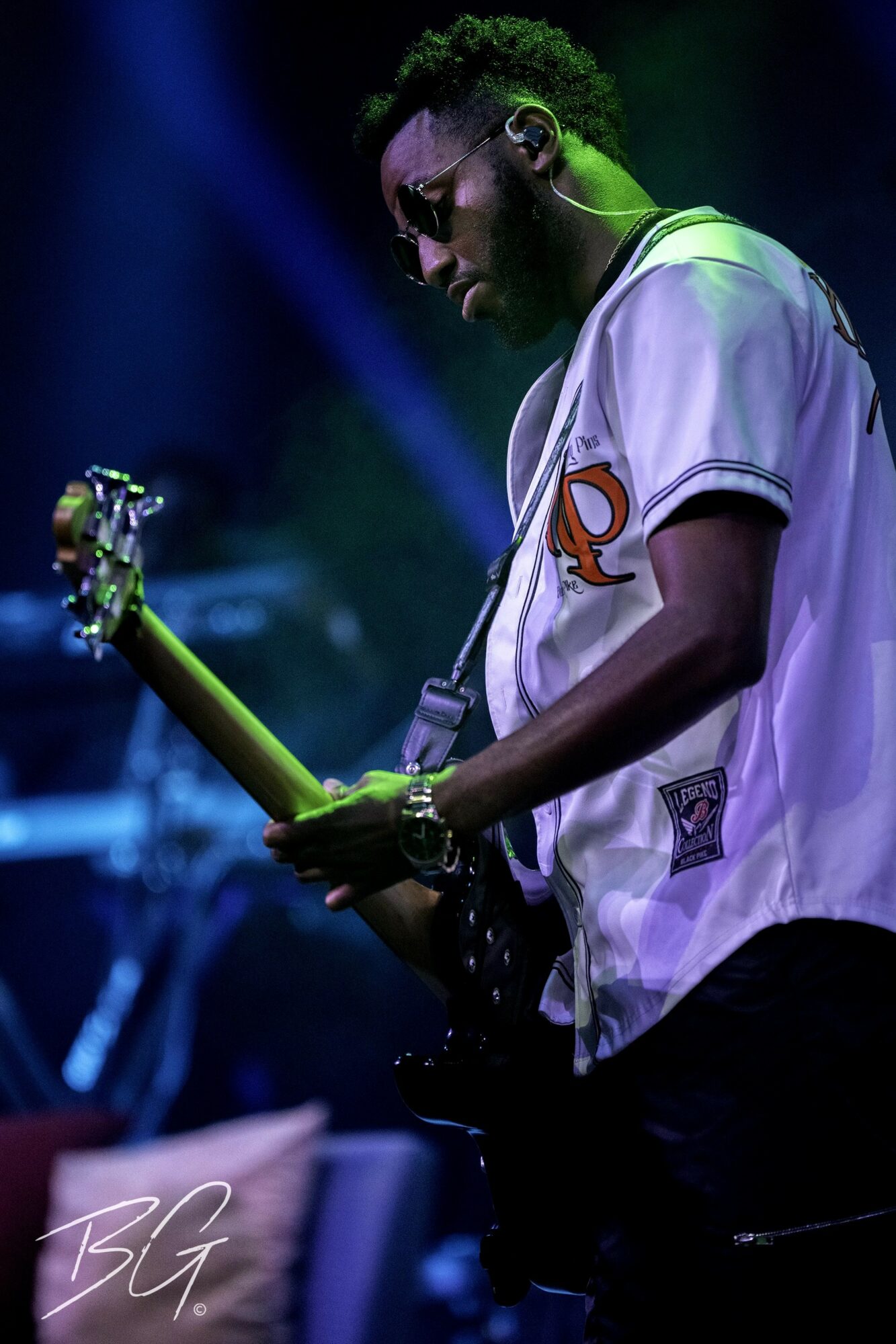 Musician playing guitar on stage with colorful lighting, wearing a white shirt and glasses.