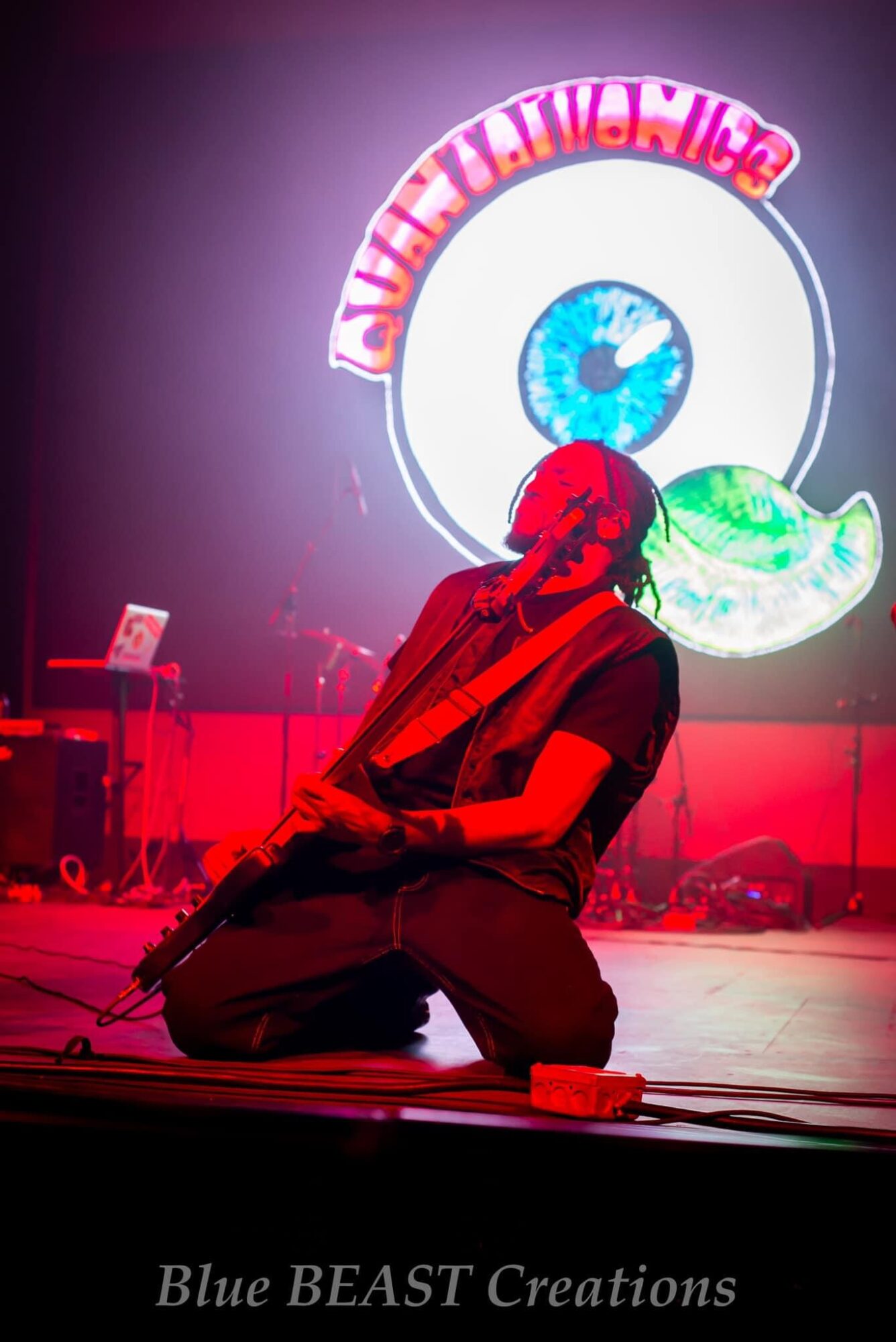 Person kneeling on stage with a large eye and colorful logo behind them, stage lighting in red and purple hues.