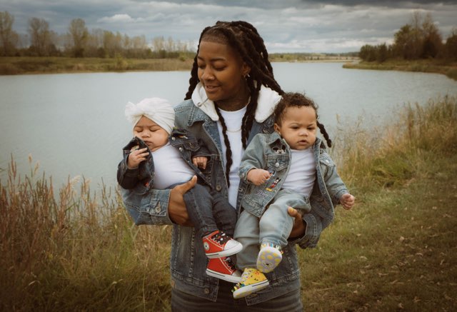Woman holding two children outdoors near a lake, with trees and cloudy sky in background.