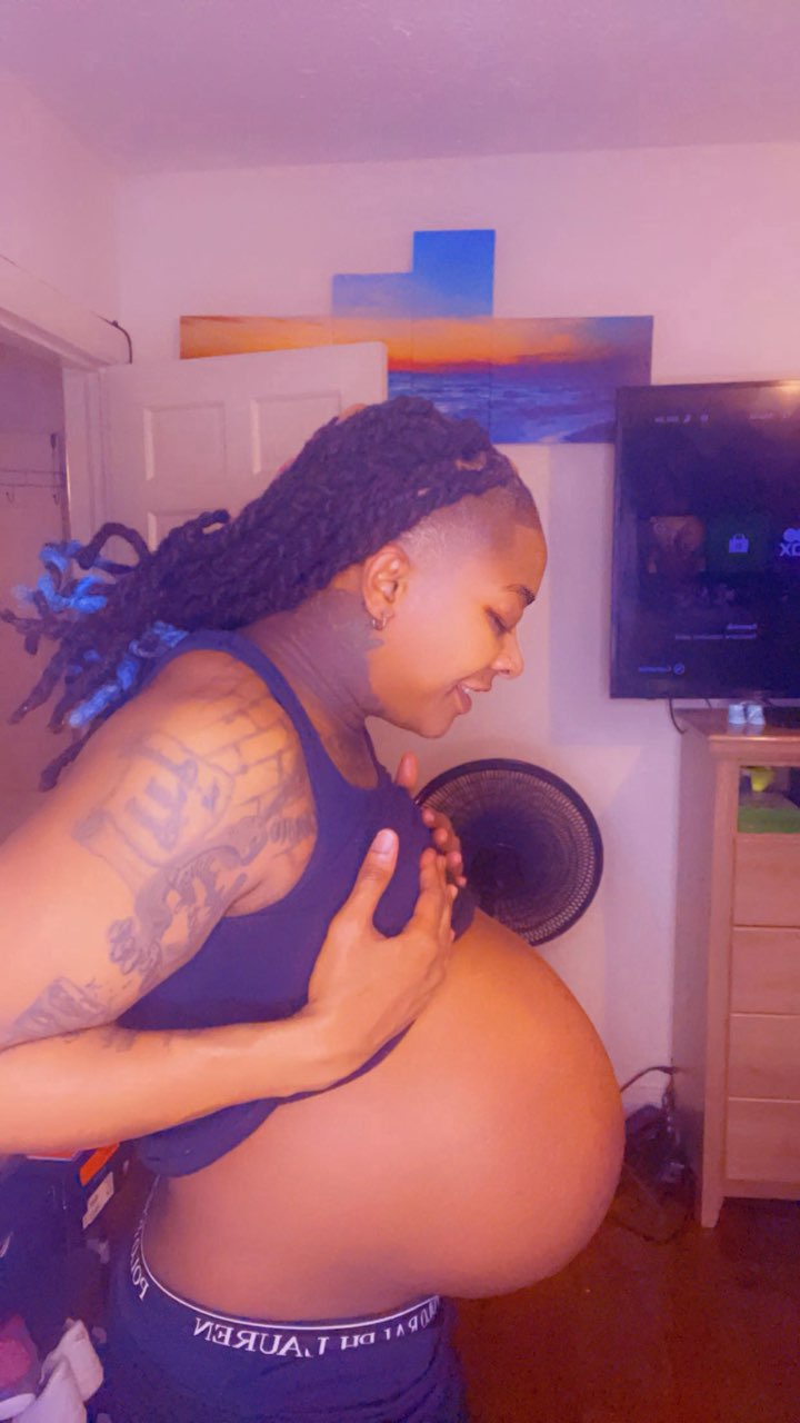 Pregnant woman with dreadlocks and tattoos, holding her belly in a room with a TV and colorful artwork.