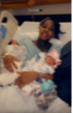 Person in hospital bed holding a newborn baby, smiling, with medical equipment nearby.
