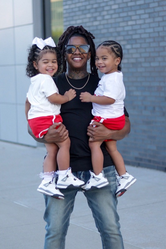 Woman holding two children outdoors, smiling, with a brick wall background.