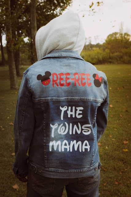 Person wearing a denim jacket with patches and text, hood up, outdoors in a park with trees and grass.