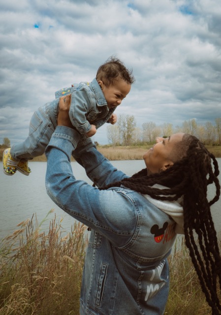 Woman with long dreadlocks lifting a smiling child outdoors near a body of water, cloudy sky above.