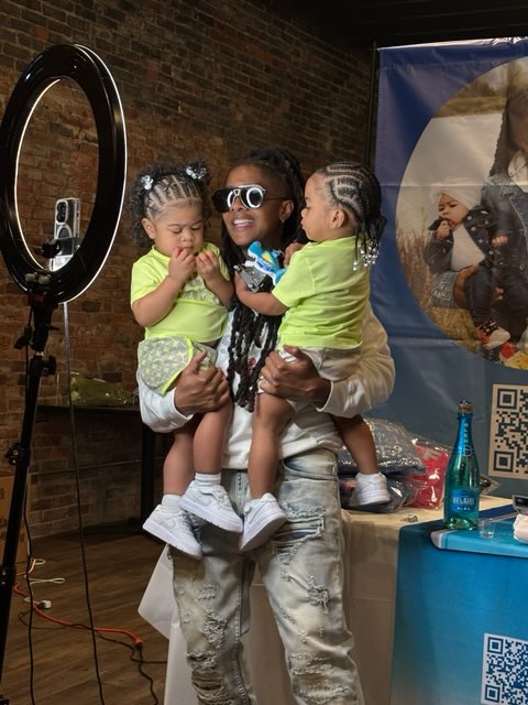 Person with sunglasses holding two children in front of a photo booth, brick wall background, children wearing green shirts.