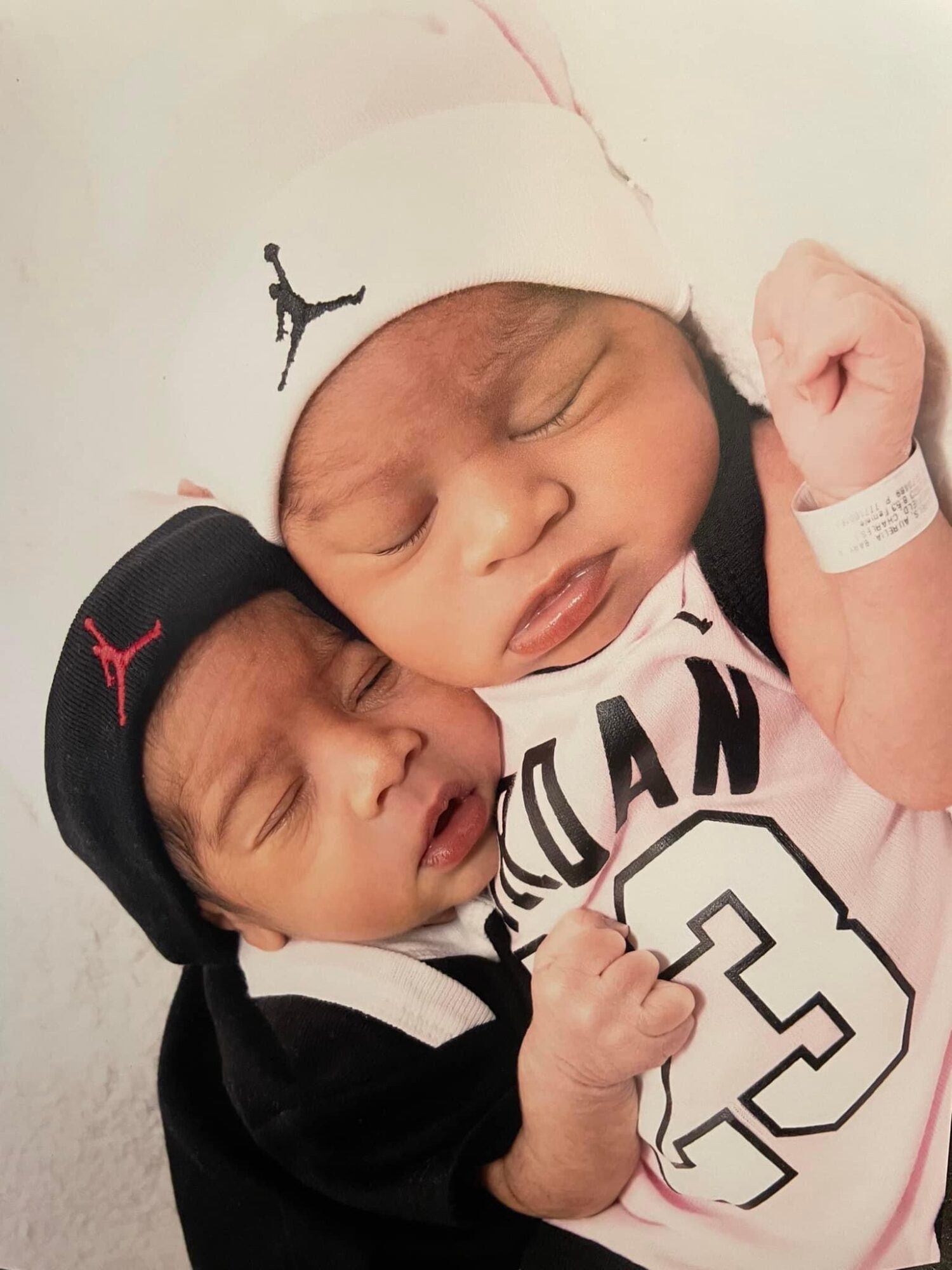 Two newborn babies lying close together, one wearing a black hat and the other a white hat, both with closed eyes.