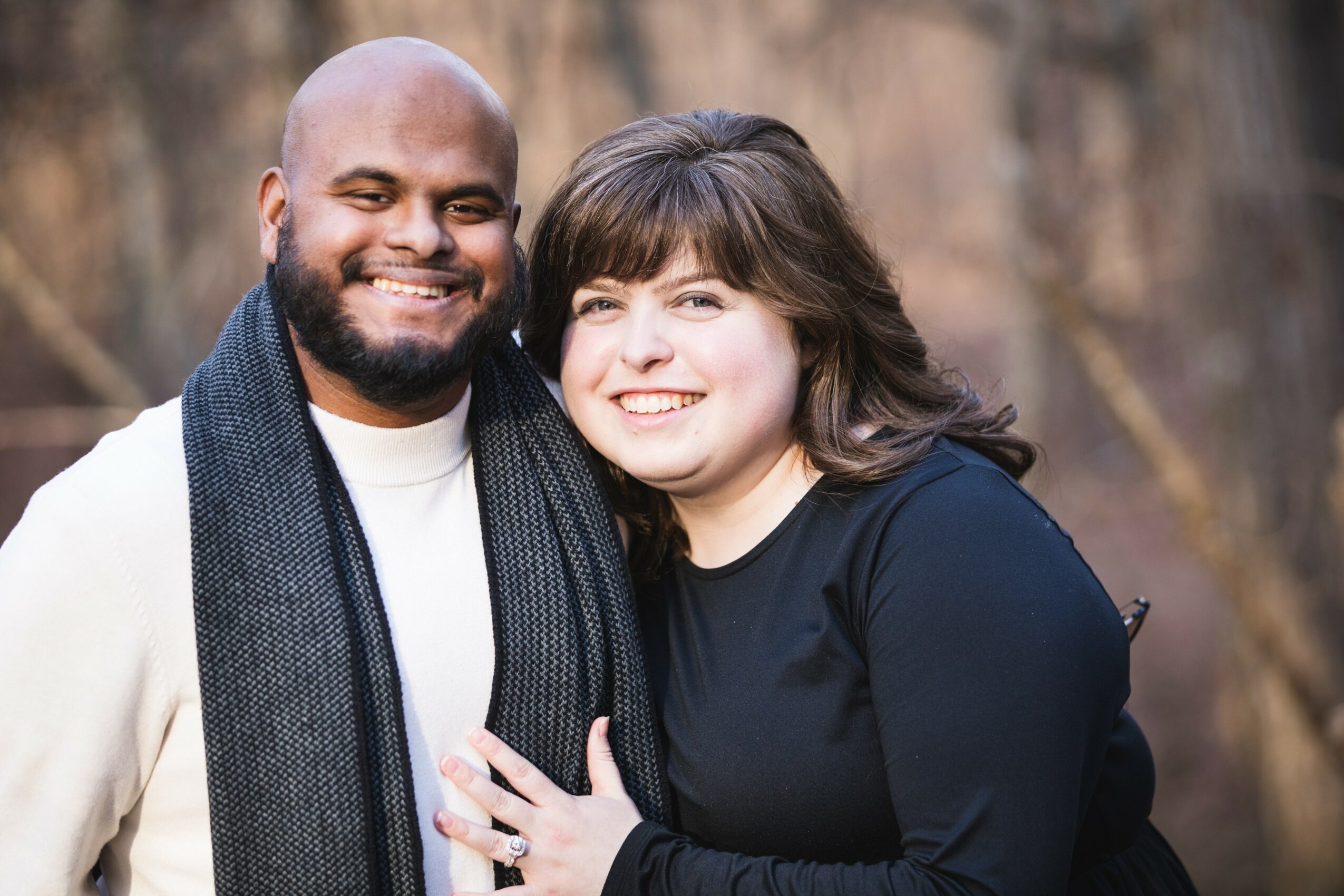 A smiling man and woman stand outdoors, close together, with trees in the background.