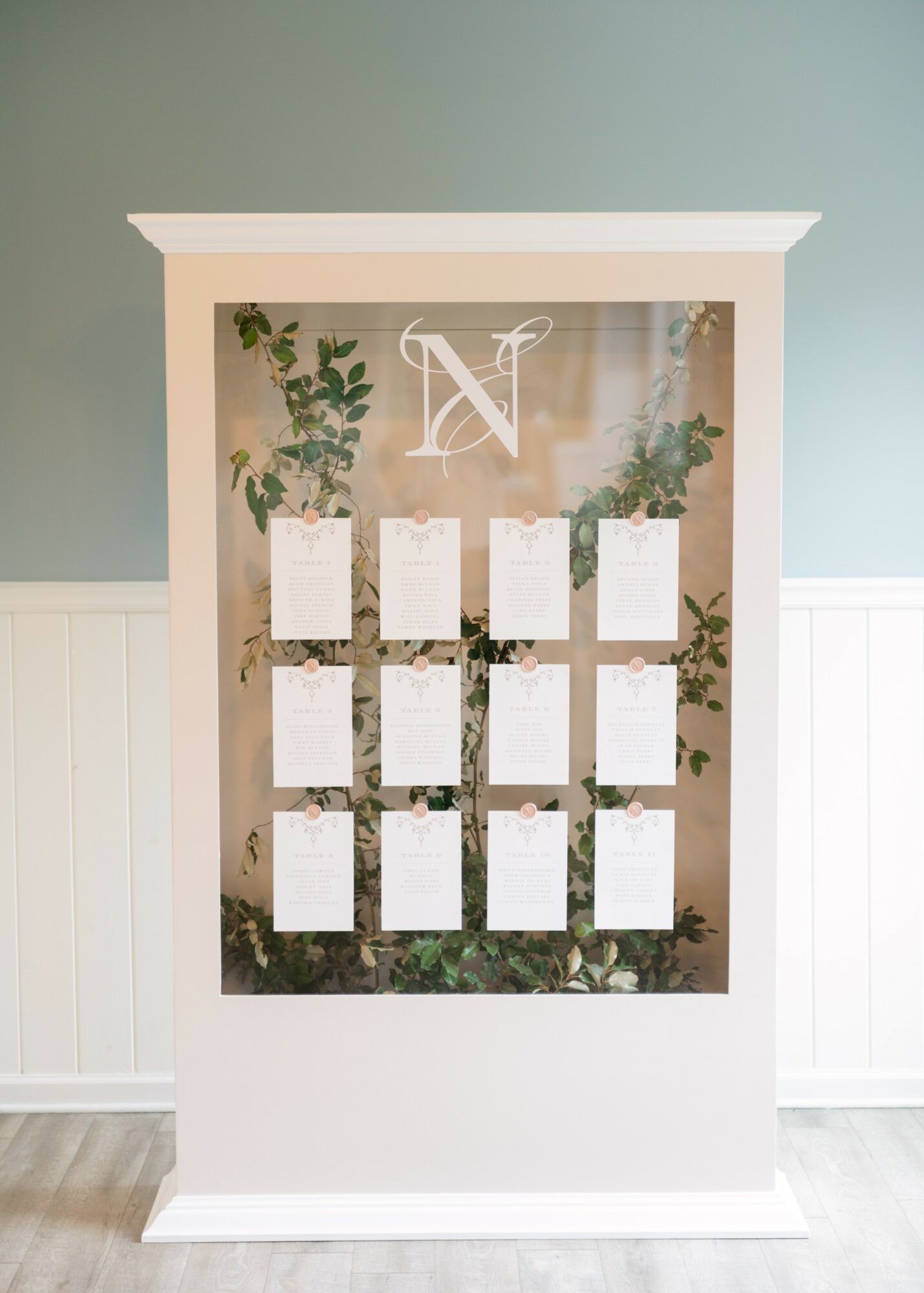 White display case with glass front, containing nine white cards and green foliage, against a light blue wall.