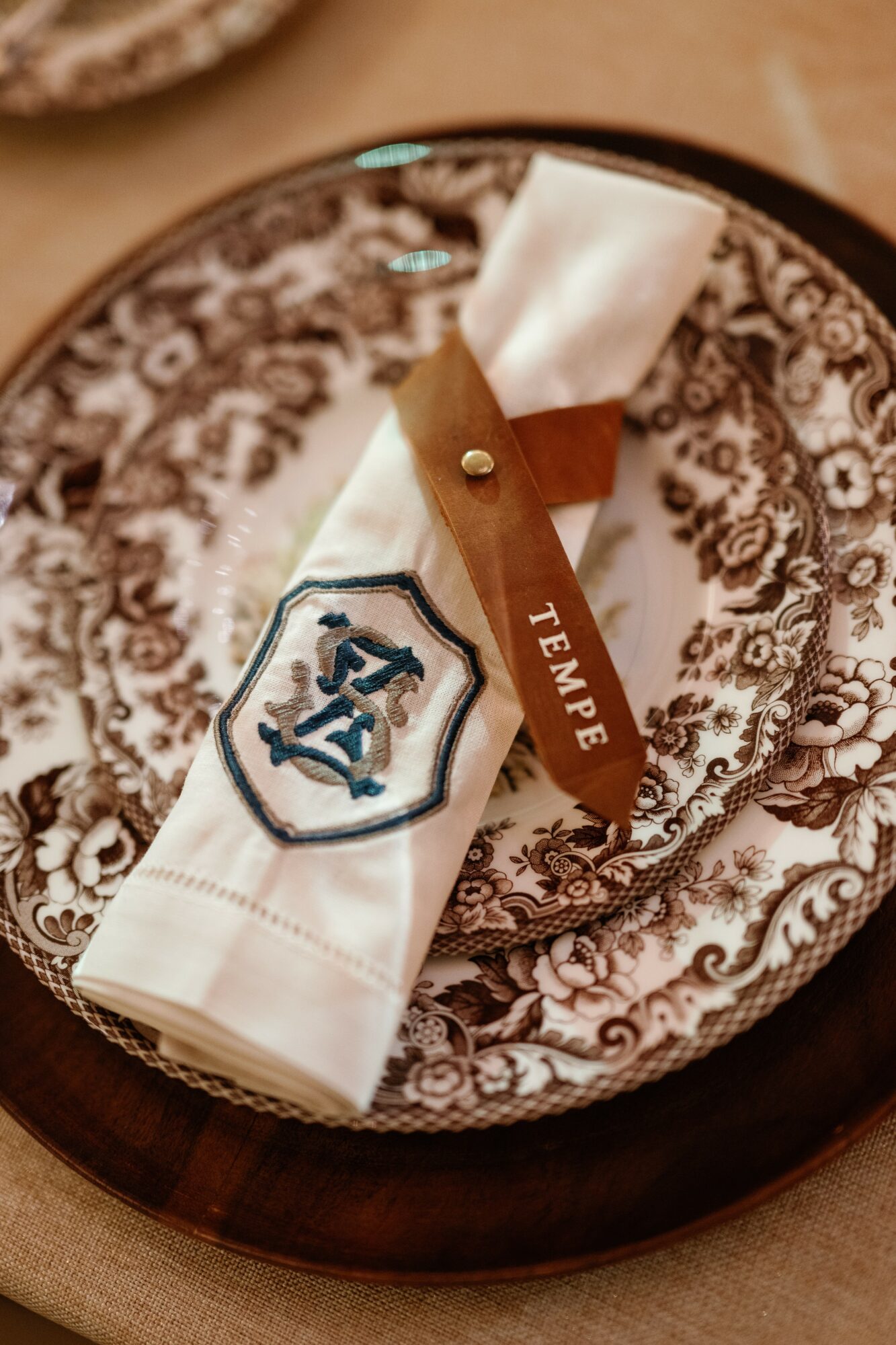 Decorative plate with a napkin, a leather tag labeled 'TEMPLE,' and a cloth with a crest, on a wooden table.