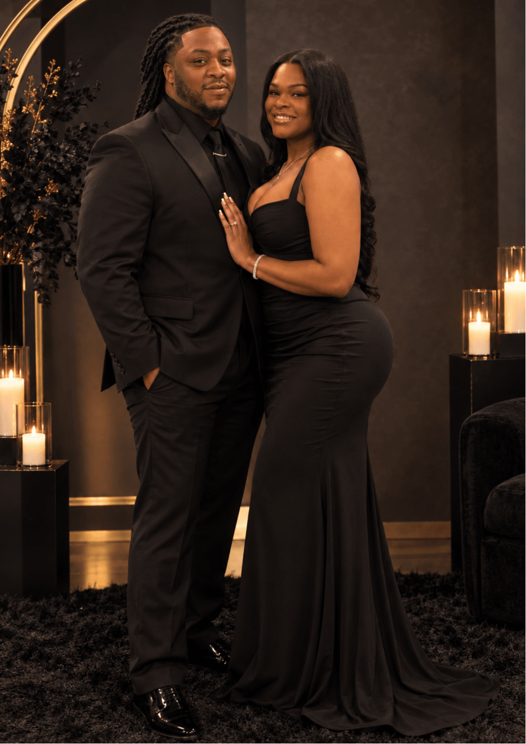 A man and woman in formal attire pose together indoors, with candles and dark decor in the background.