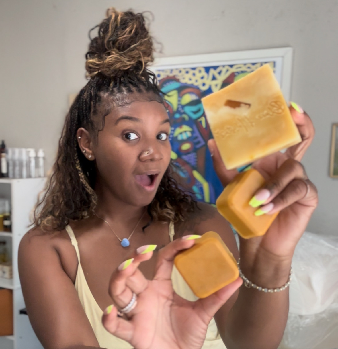 Young woman with curly hair holding two bars of soap, smiling, in a room with artwork on the wall.