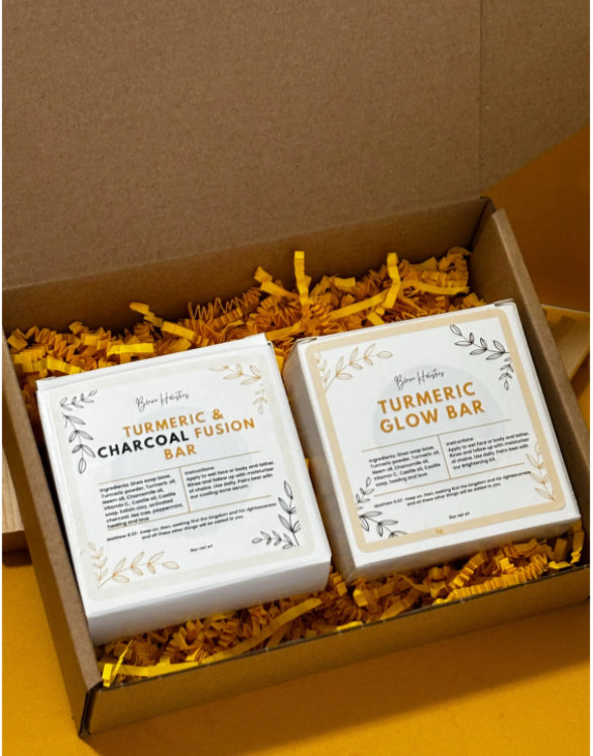 Two boxes labeled turmeric glow bar and turmeric charcoal fusion bar inside a gift box with yellow shredded paper, viewed from above.