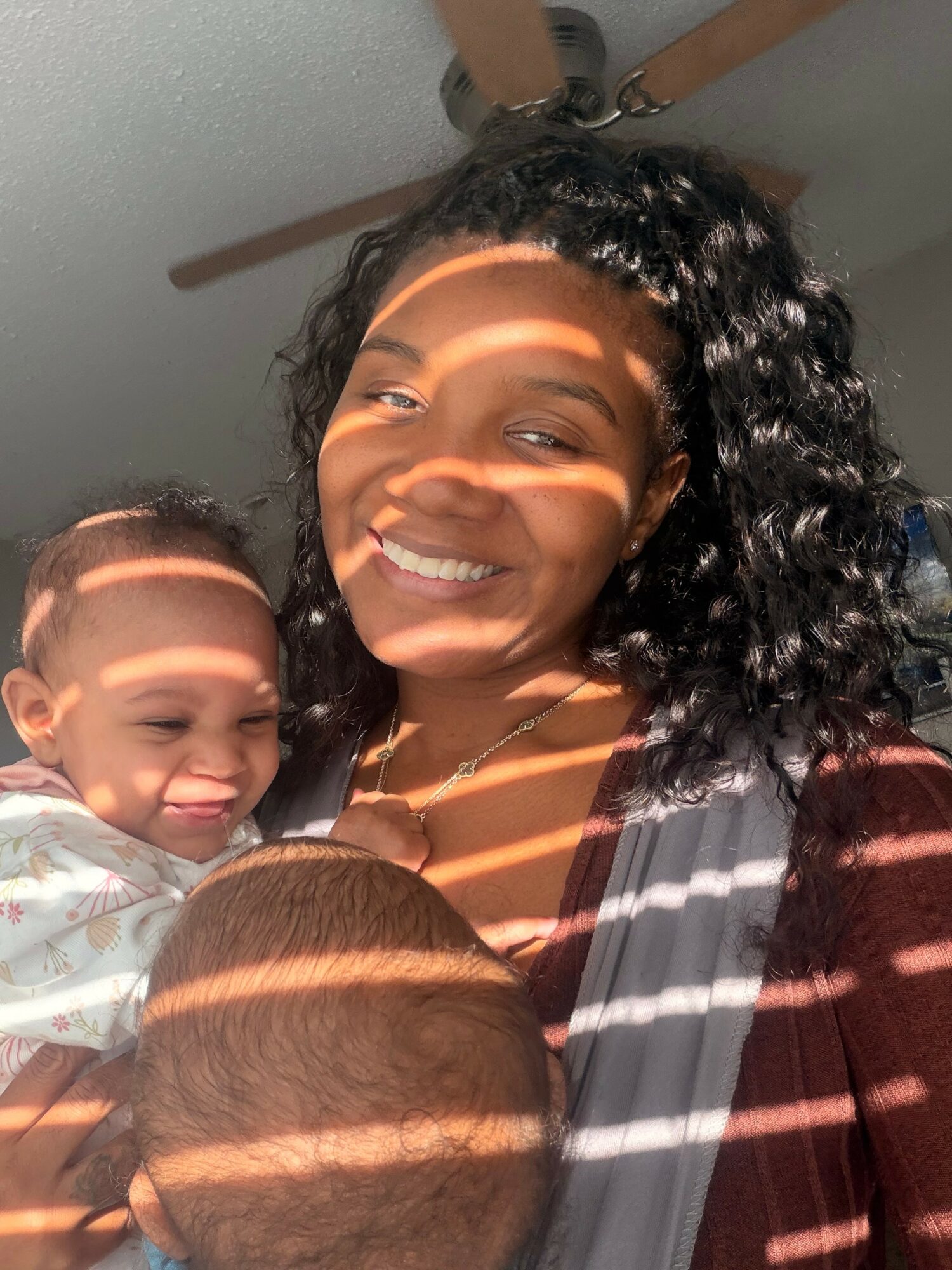 Smiling woman holding two children, with sunlight and shadows on their faces, ceiling fan above, in a room.