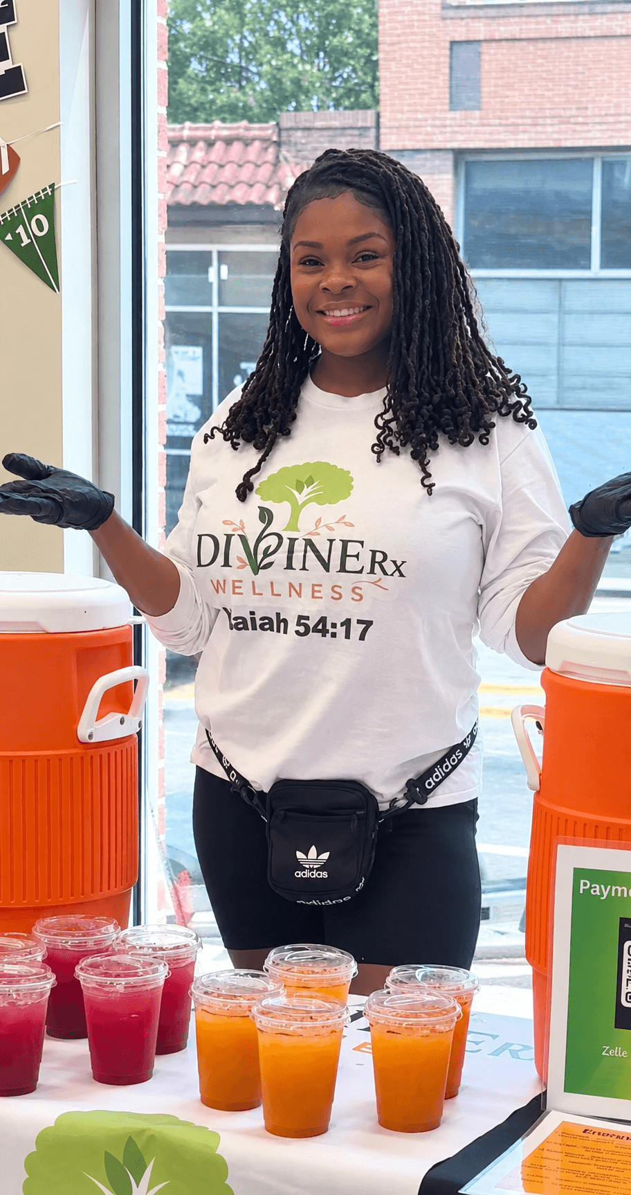 Smiling woman with long curly hair wearing a white T-shirt and black gloves, standing behind juice cups inside a store.