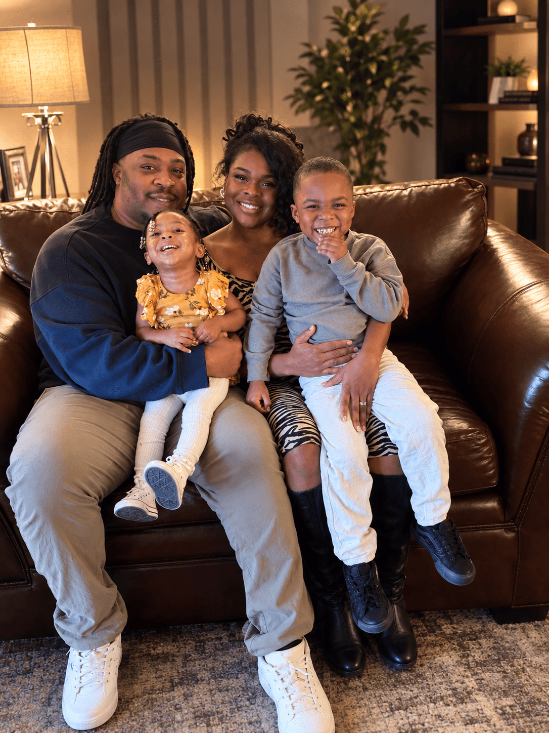 Family of four sitting on a brown leather couch, smiling, in a cozy living room with a lamp and plants.