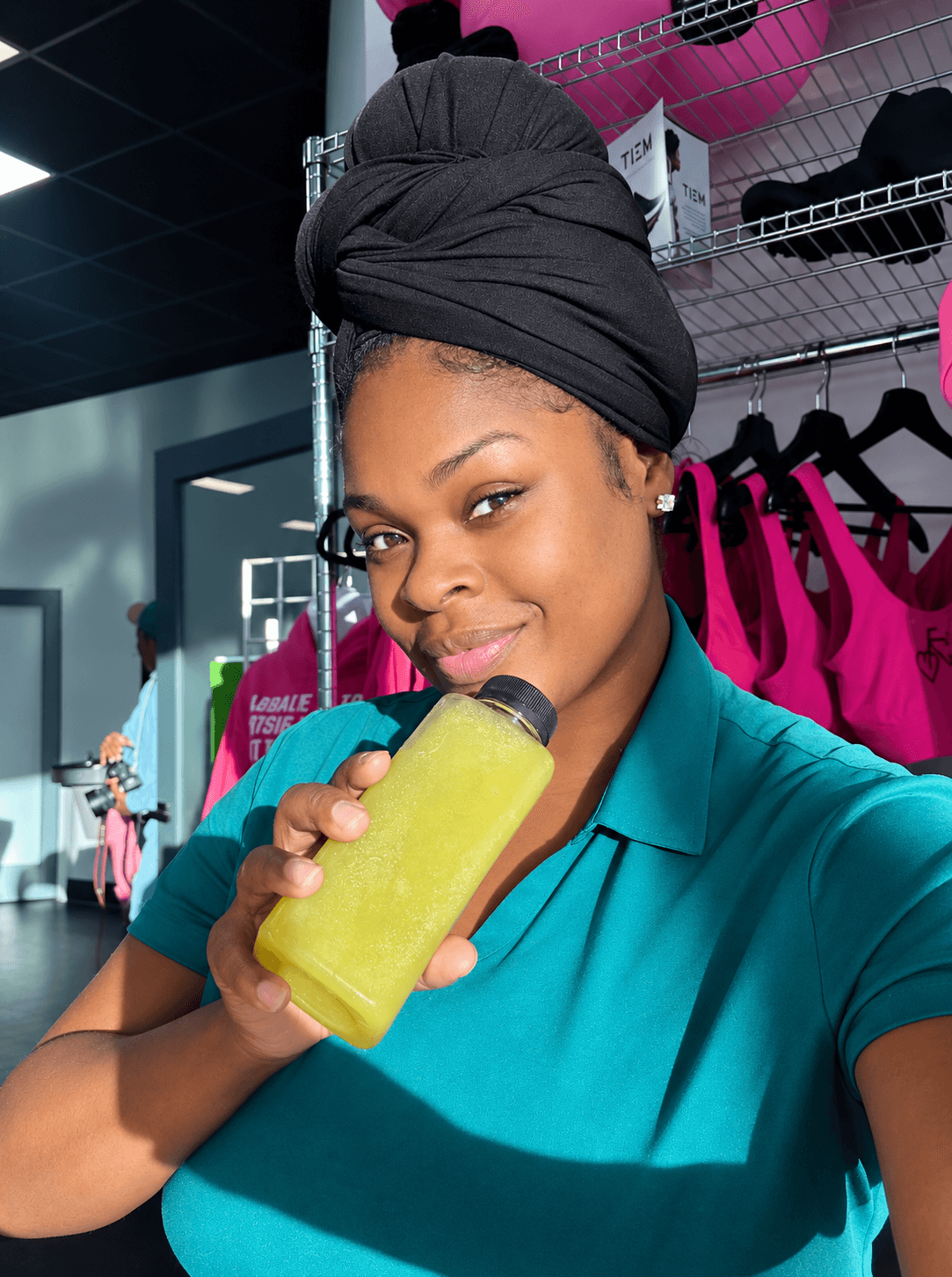 Young woman with a black headwrap drinking a yellow smoothie in a store with pink and black clothing displays.