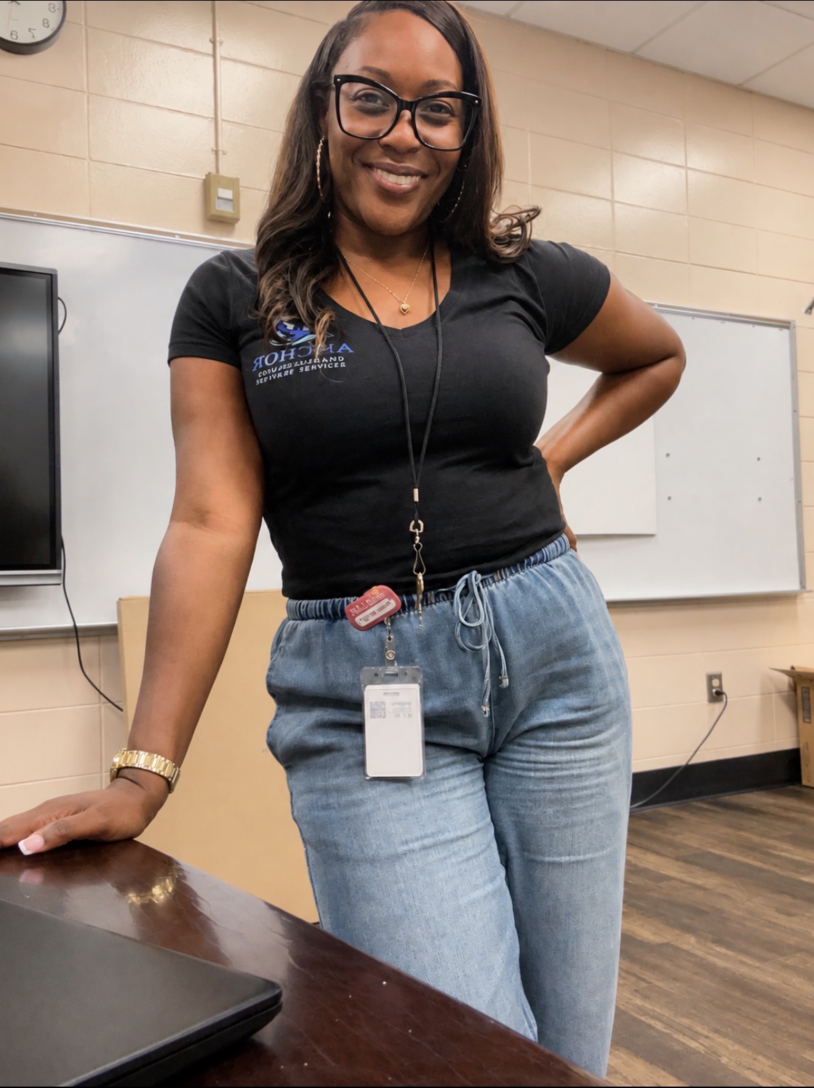 Smiling woman with glasses and long hair, wearing a black t-shirt and jeans, standing in a classroom.