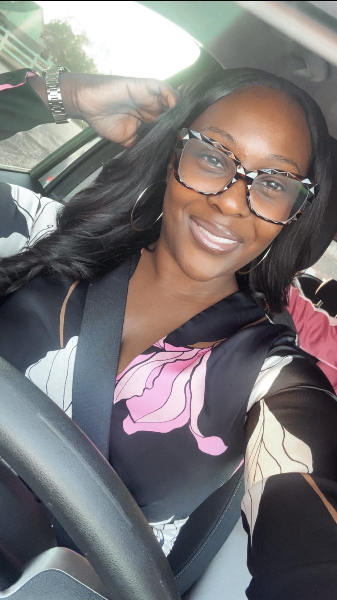 Woman with glasses smiling in car, wearing a colorful top and carrying a backpack.