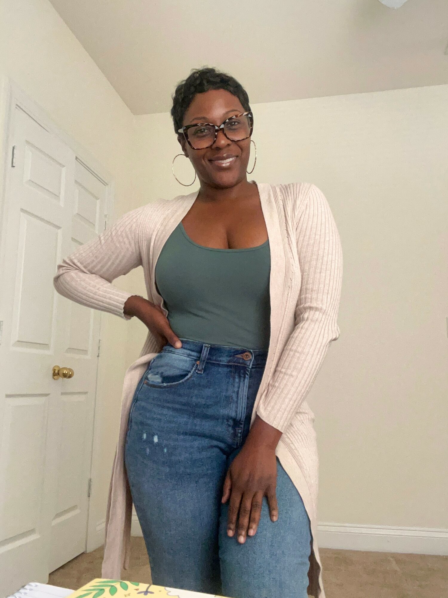 Woman with glasses and hoop earrings smiling, standing indoors with hand on hip, wearing a teal top and beige cardigan.