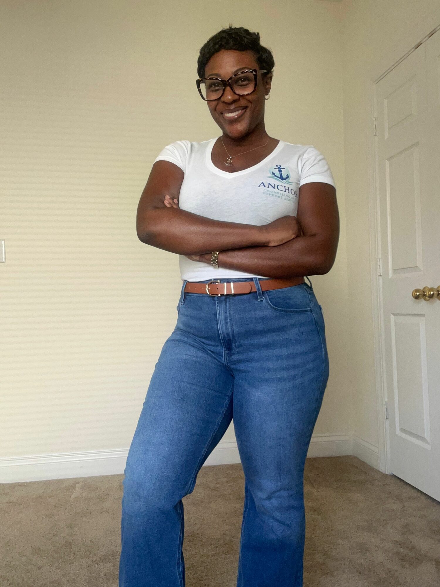 Person standing with arms crossed, wearing glasses, white t-shirt, and blue jeans, smiling indoors.