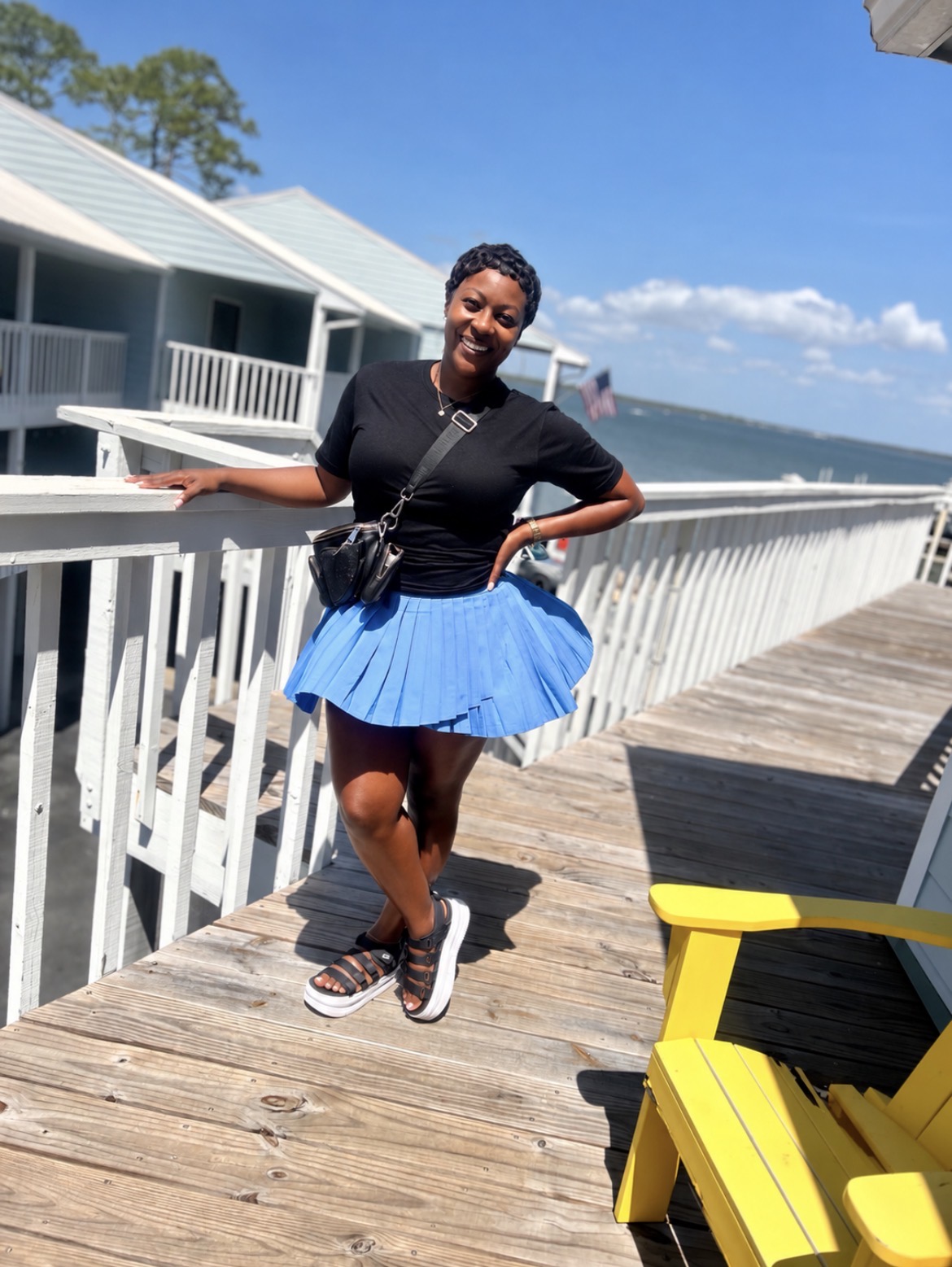 Woman standing on a wooden deck near water, smiling, wearing a black top and blue skirt, with a yellow chair nearby.
