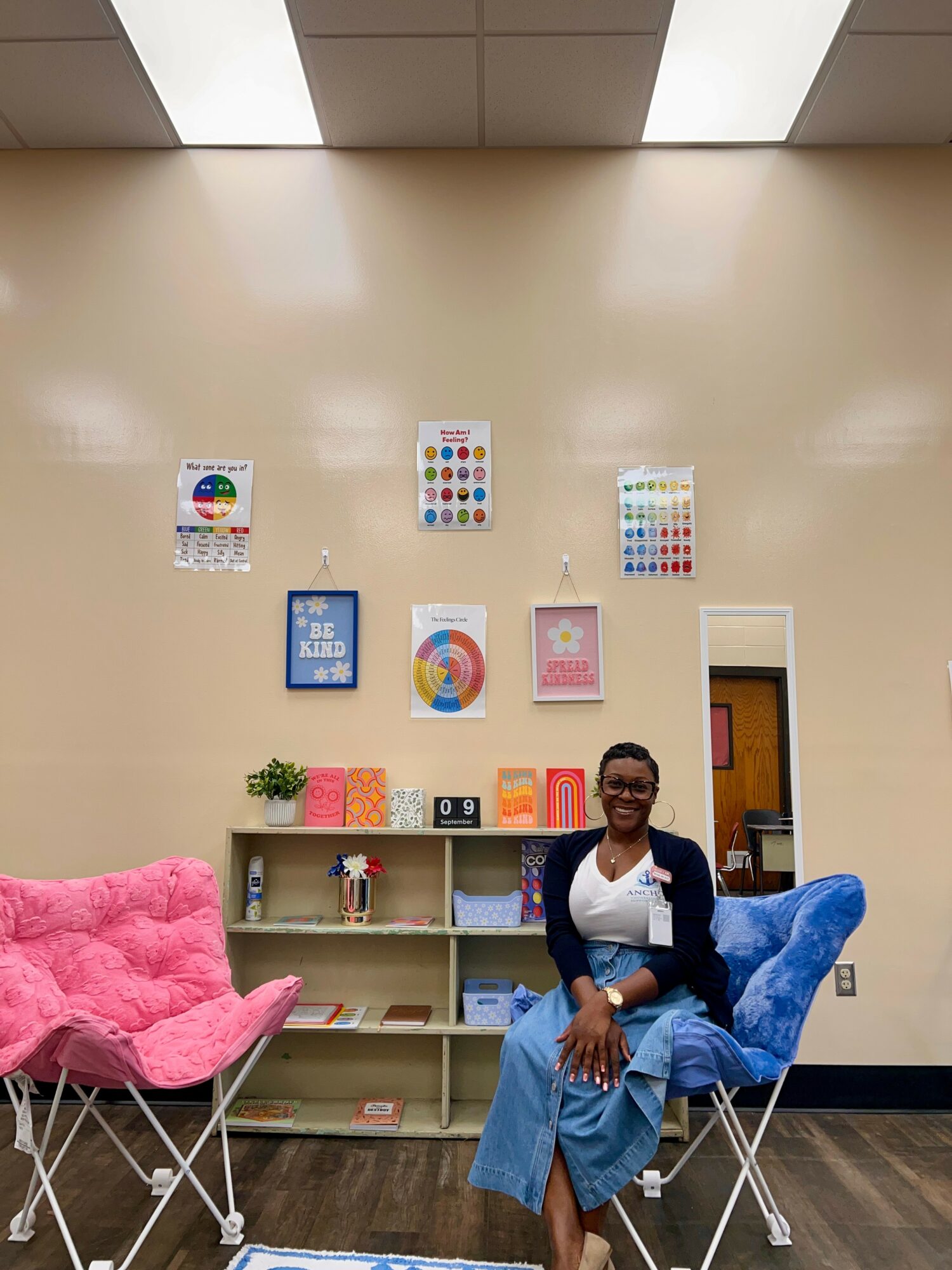 Woman sitting on a blue chair in a room with a beige wall decorated with colorful posters, pink chair nearby, bookshelf behind her.