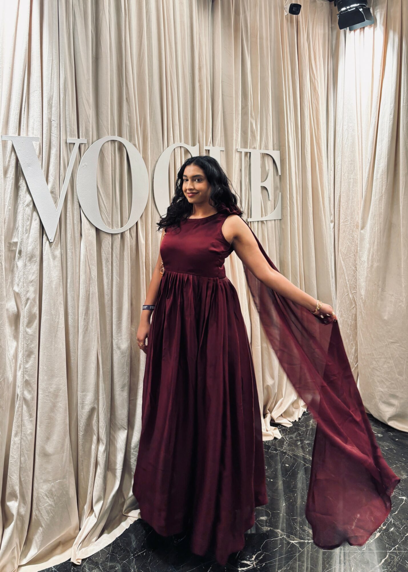 Woman in a long burgundy dress standing in front of large white letters spelling 'LOVE' with beige curtains behind her.