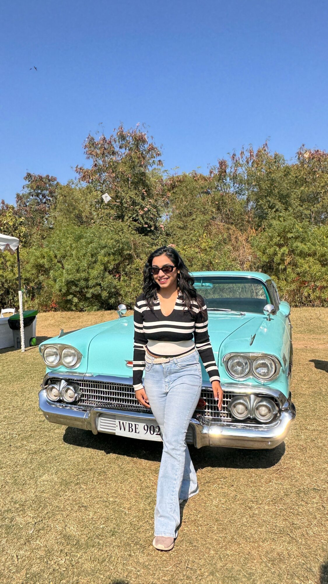 Woman in sunglasses and striped shirt standing in front of a vintage blue car outdoors, trees and blue sky in background.