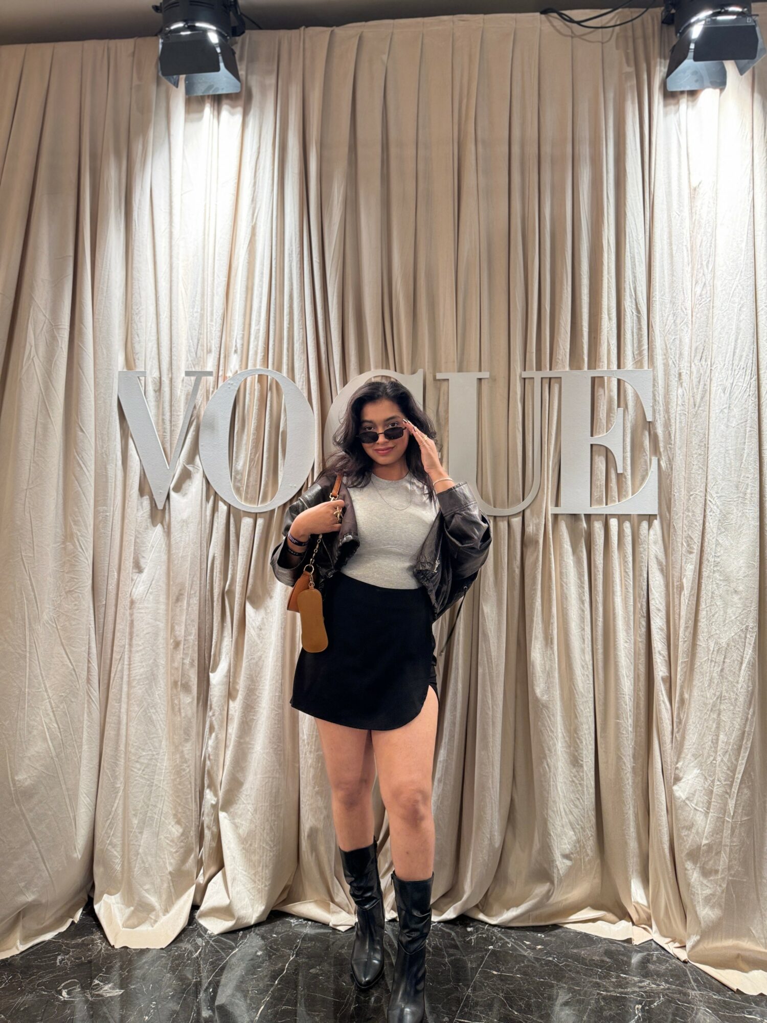 Woman with sunglasses and a black jacket standing in front of a Vogue sign, beige curtains backdrop.