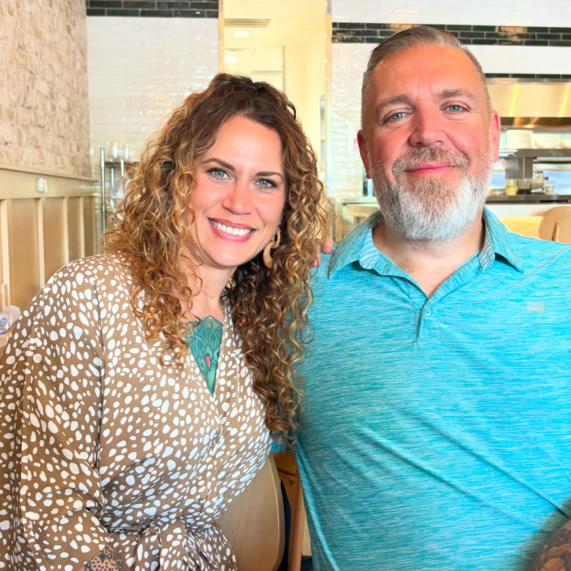Two people smiling, woman with curly hair and a man with a beard, in a bright indoor setting.